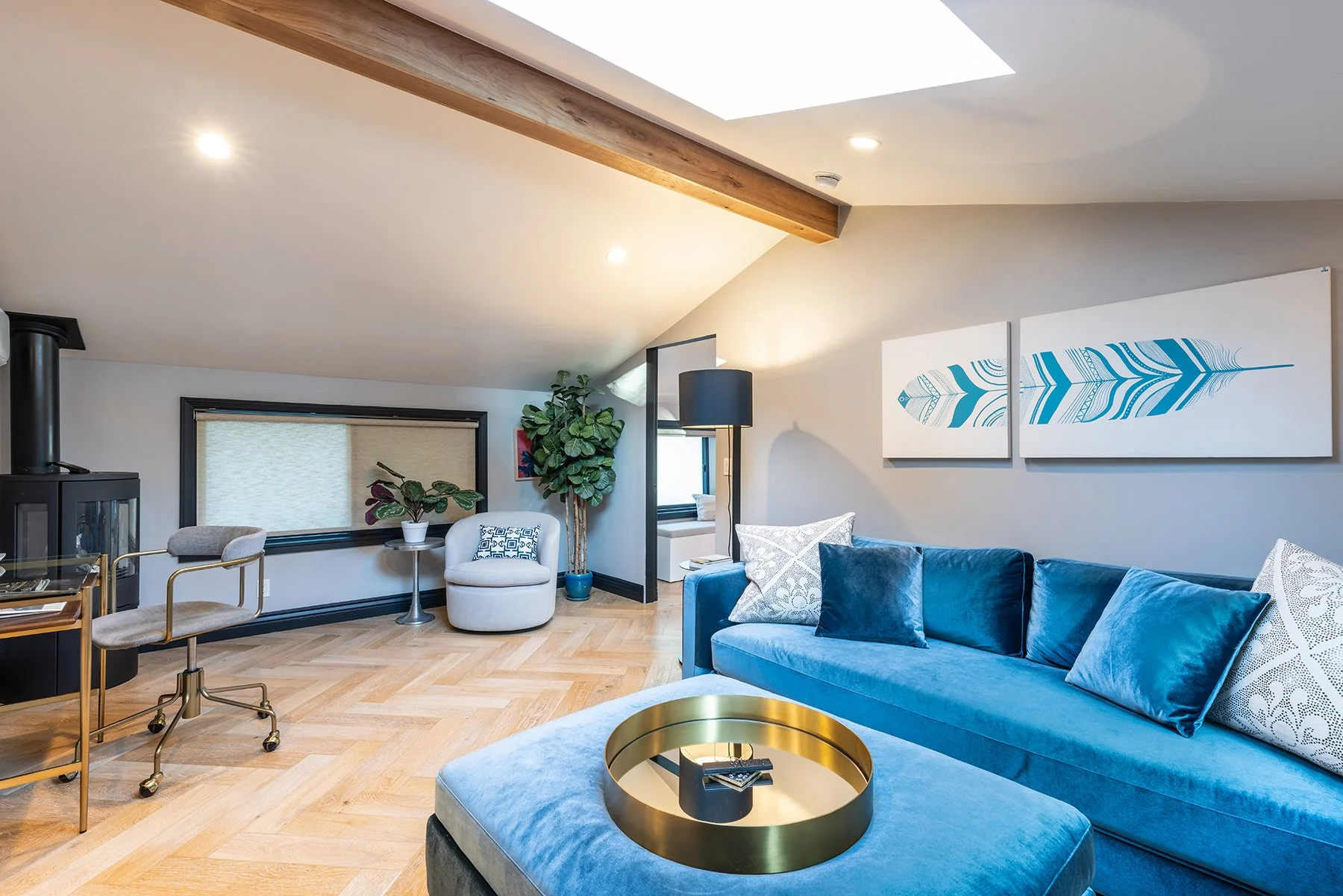 Modern living room with blue velvet sofa, white and patterned throw pillows, gold round coffee table, large artwork of a blue feather, beige armchair, green plants, black fireplace, window with beige blind, and wooden floor.
