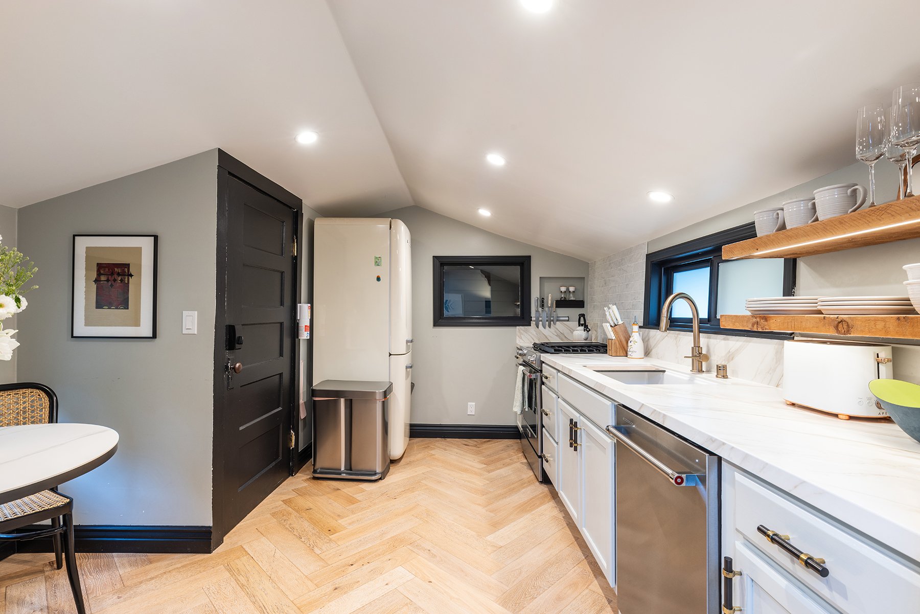 Modern kitchen with white cabinetry, marble countertops, a stainless steel dishwasher, and a black-framed window above the sink. There are open wooden shelves with white dishes and glasses, and a retro-style refrigerator next to a black door. A small