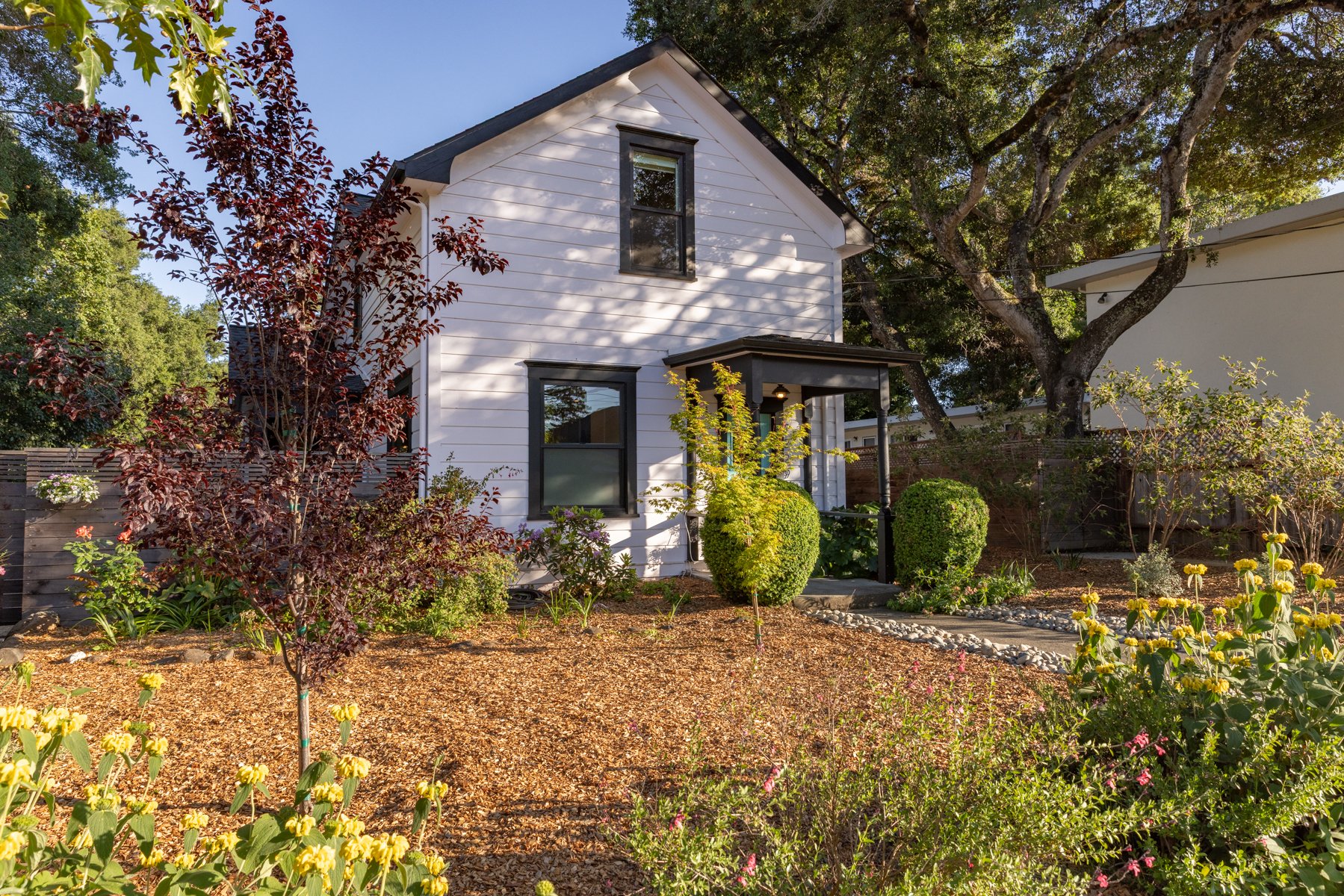 White house with black trim surrounded by a garden with various plants, trees, and flowers on a sunny day.
