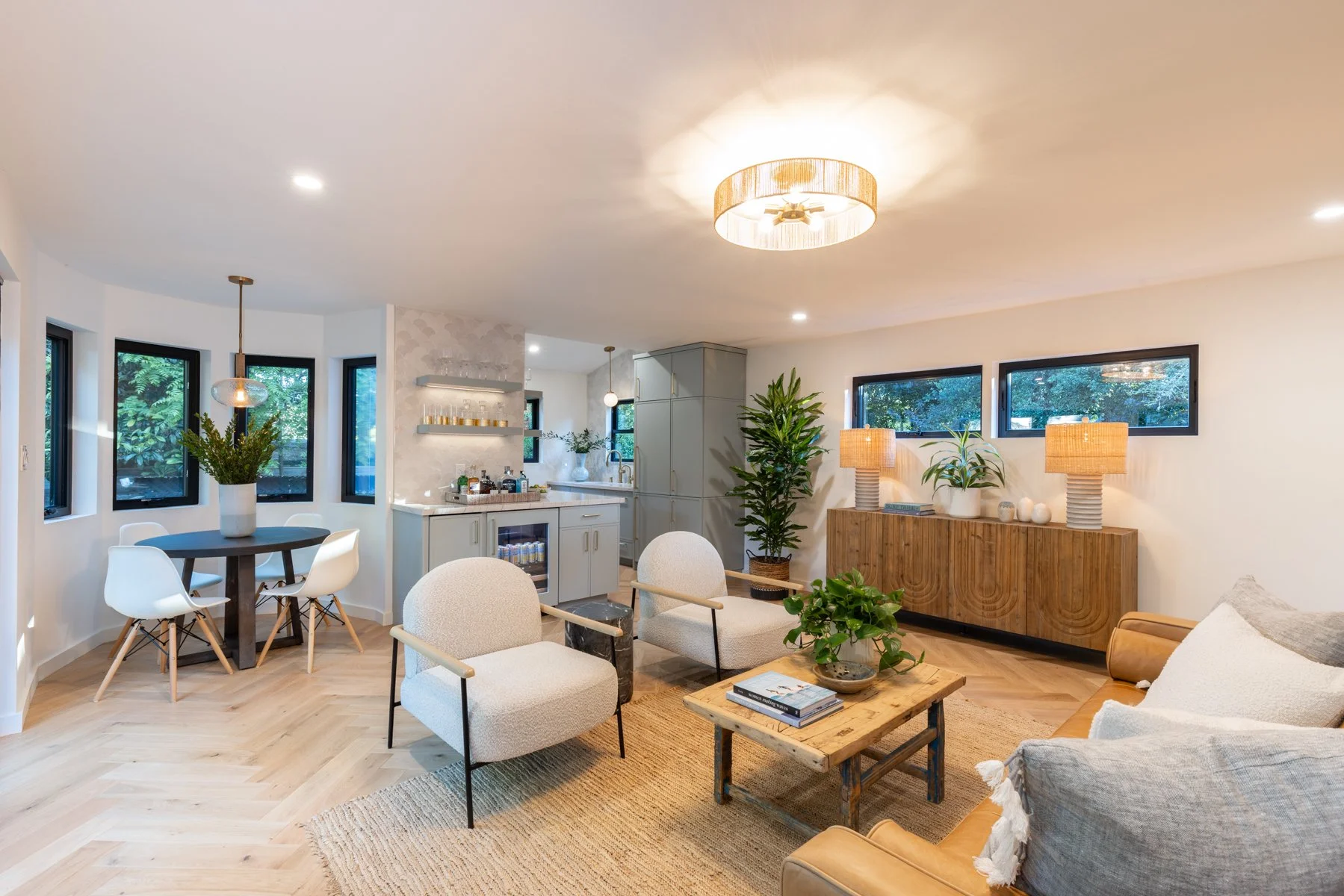 A spacious, modern living room with natural light streaming through multiple windows. It features neutral tones, wooden accents, a dining area with a round table and white chairs, a cozy seating area with armchairs and a coffee table, and a small bar