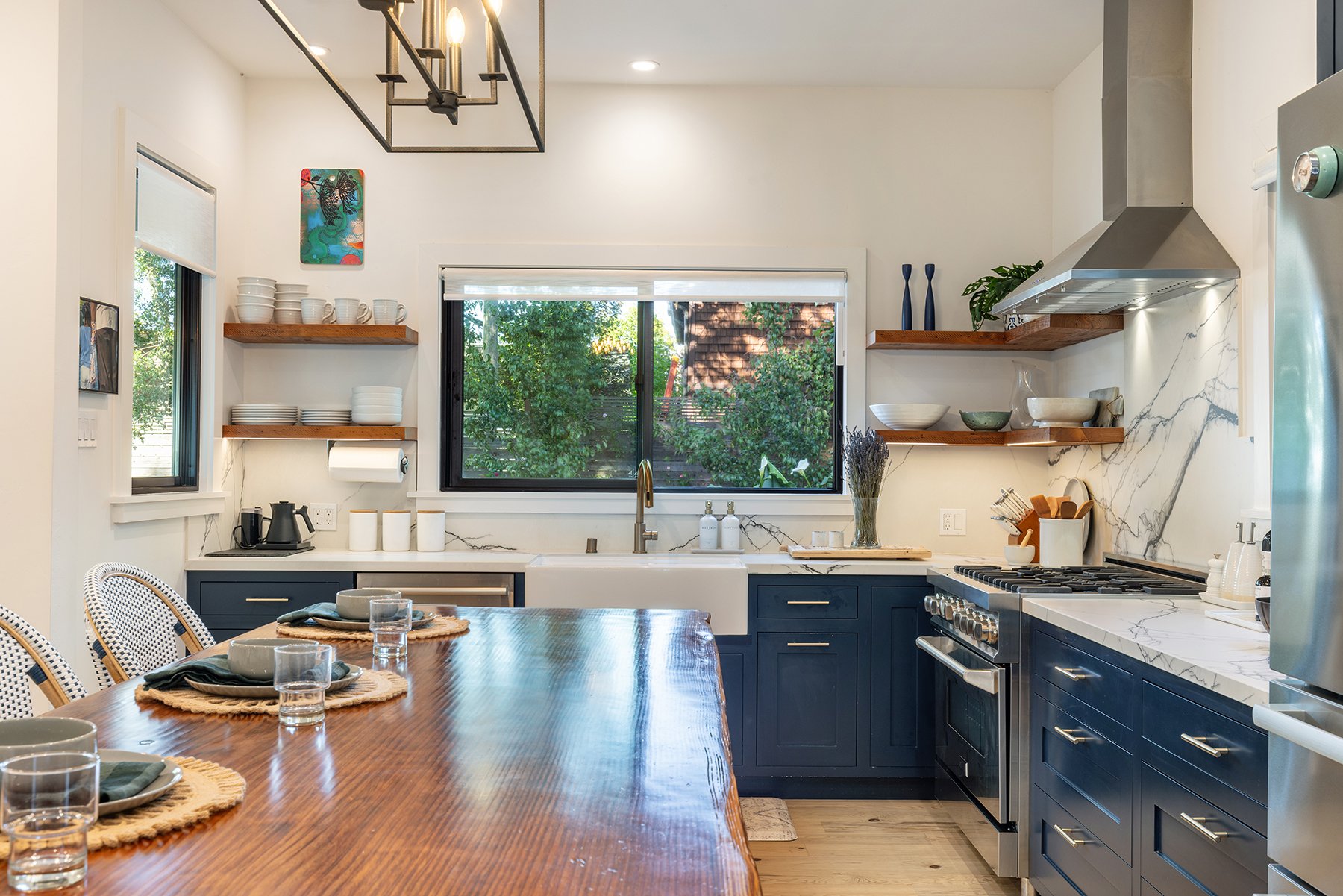 A modern kitchen with navy blue cabinets, marble countertops, open wooden shelves with bowls and decorative vases, a large window overlooking trees, and a wooden dining table set with plates, glasses, and napkins.