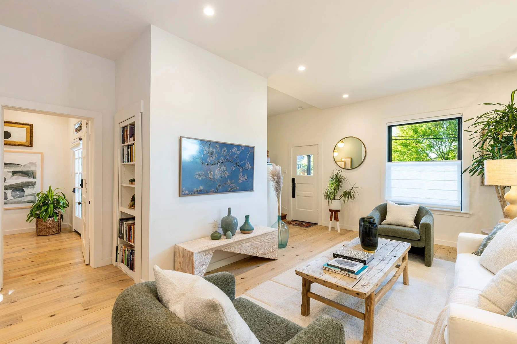 Bright living room with large window showing green trees, modern furniture including a gray armchair, white sofa, rustic wooden coffee table, decorative vases, plants, wall art, and a mirror.