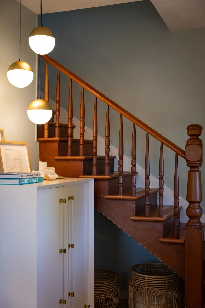 Wooden staircase with balusters and handrail next to a white cabinet with books and decorative items, hanging pendant lights, and woven baskets underneath.