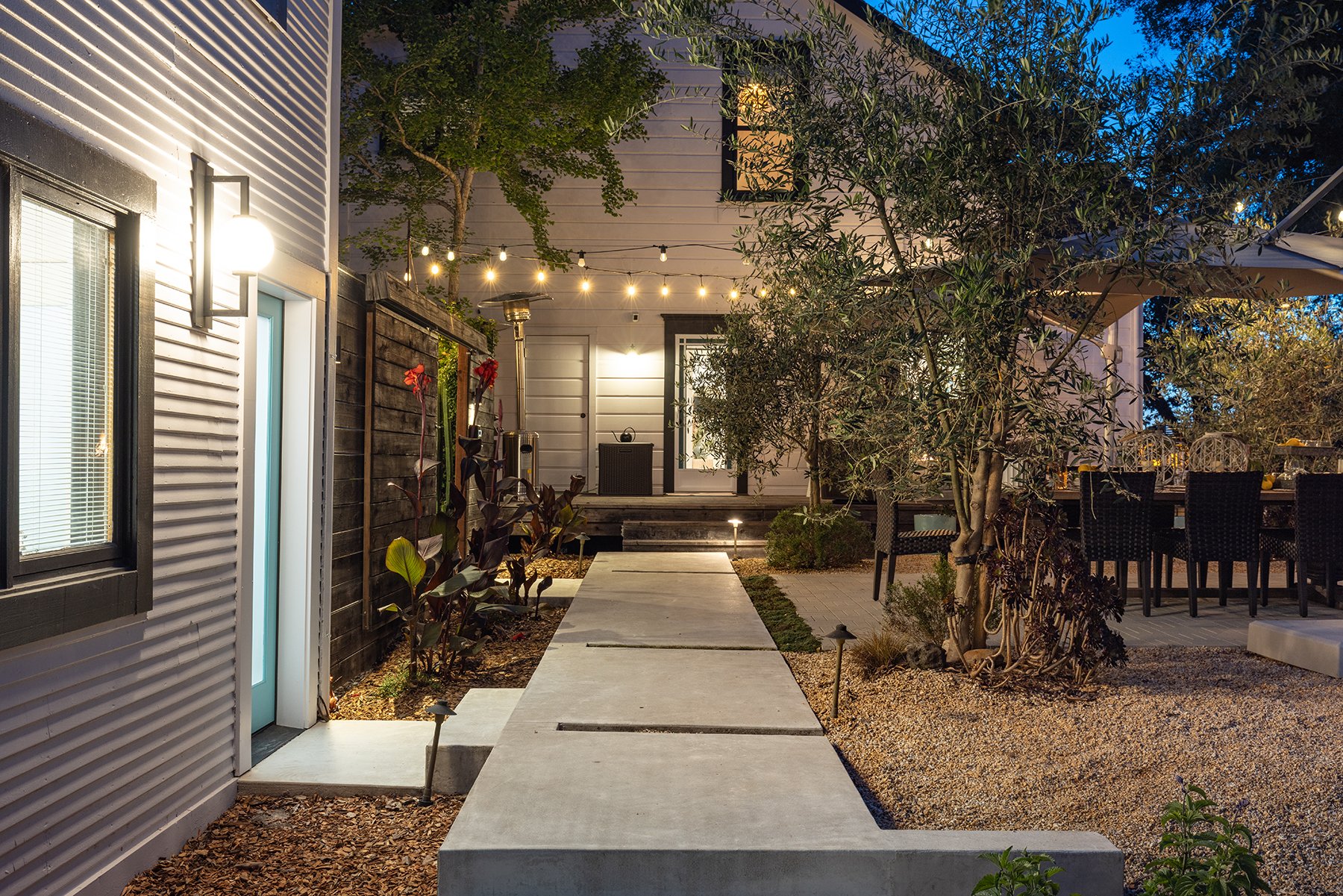 A backyard patio area illuminated with string lights at dusk, featuring a pathway made of concrete slabs leading to a house with a white exterior and black trim, a dining table with black wicker chairs, and various trees and plants.