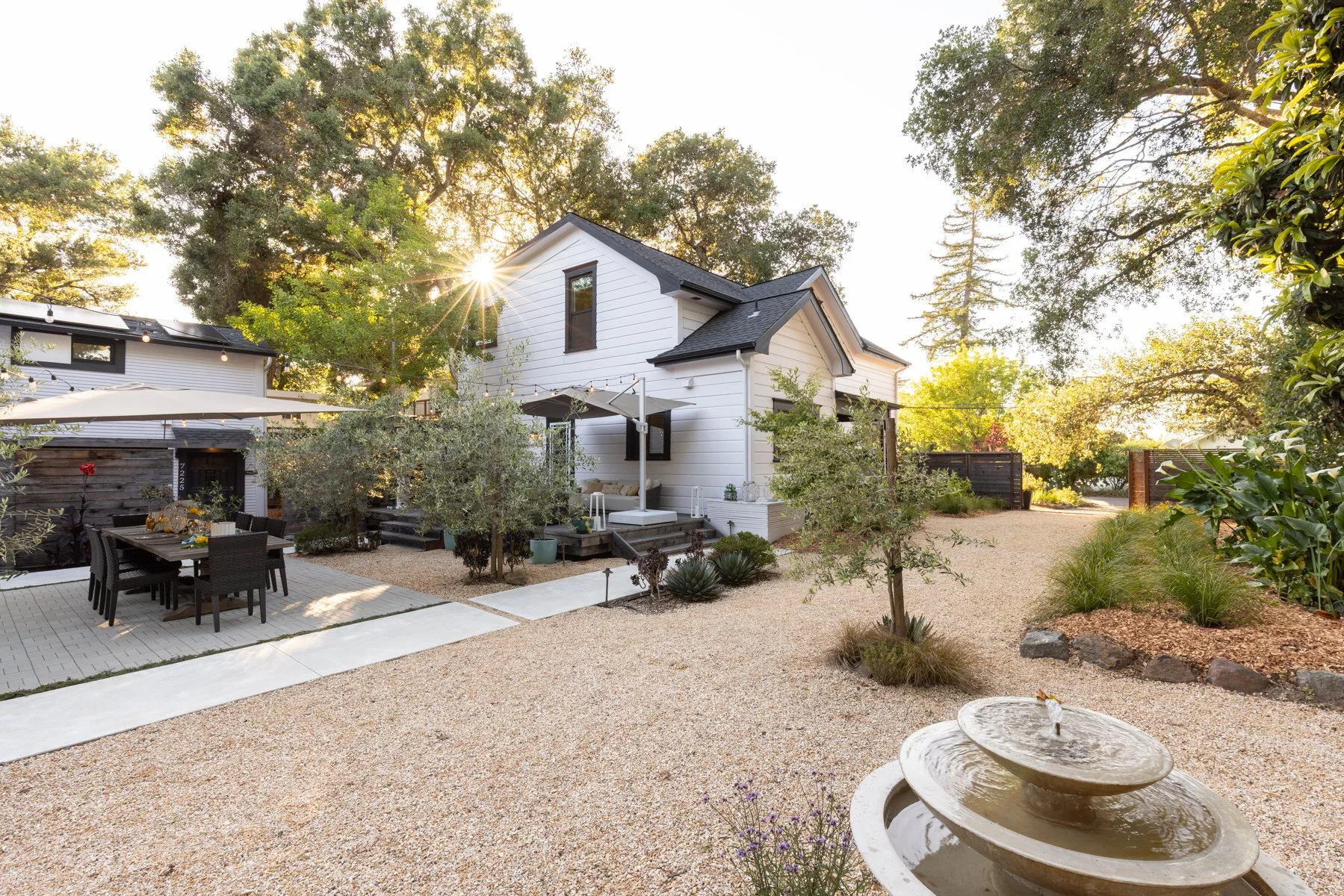 A white two-story house with black trim, surrounded by trees and a landscaped backyard. The backyard features a patio area with a dining table and chairs, umbrella, and landscaped garden with rocks and plants. The sun is setting, casting a warm glow.