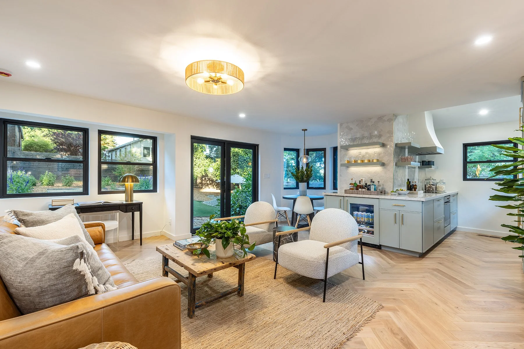 Living room with beige sofa, gray armchairs, wooden coffee table with plants, and a view of the backyard through large windows and a glass door, with a nearby kitchen area featuring white cabinets and open shelving.