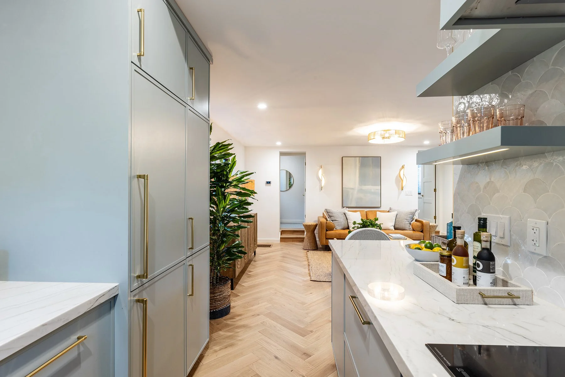 Open-concept kitchen and living room with light blue cabinetry, white marble countertops, a white tiled wall, and a cozy seating area with beige sofa and artwork.