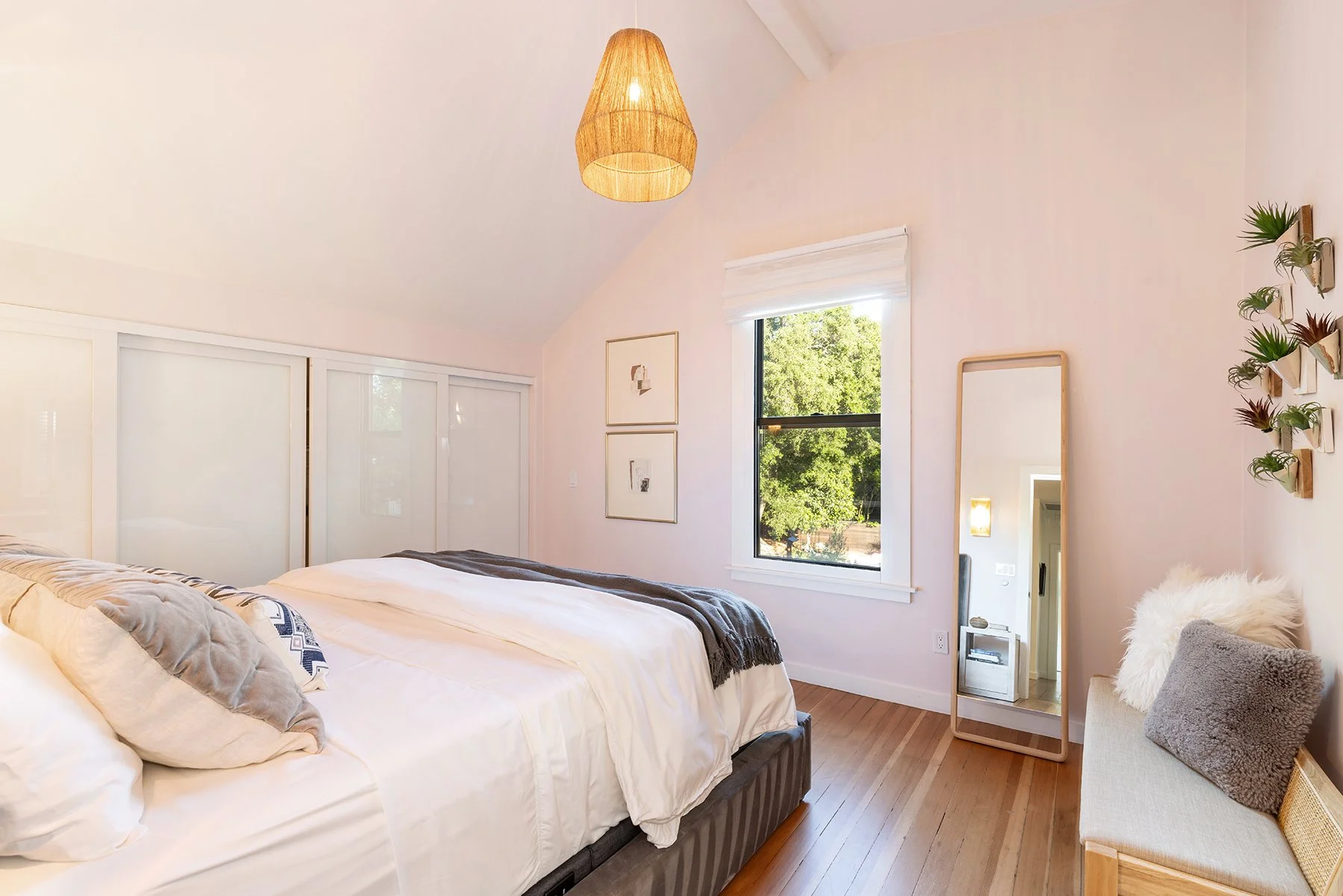 A cozy bedroom with a bed, decorative pillows, a large mirror, a window showing green trees, wall art, and a hanging light fixture.