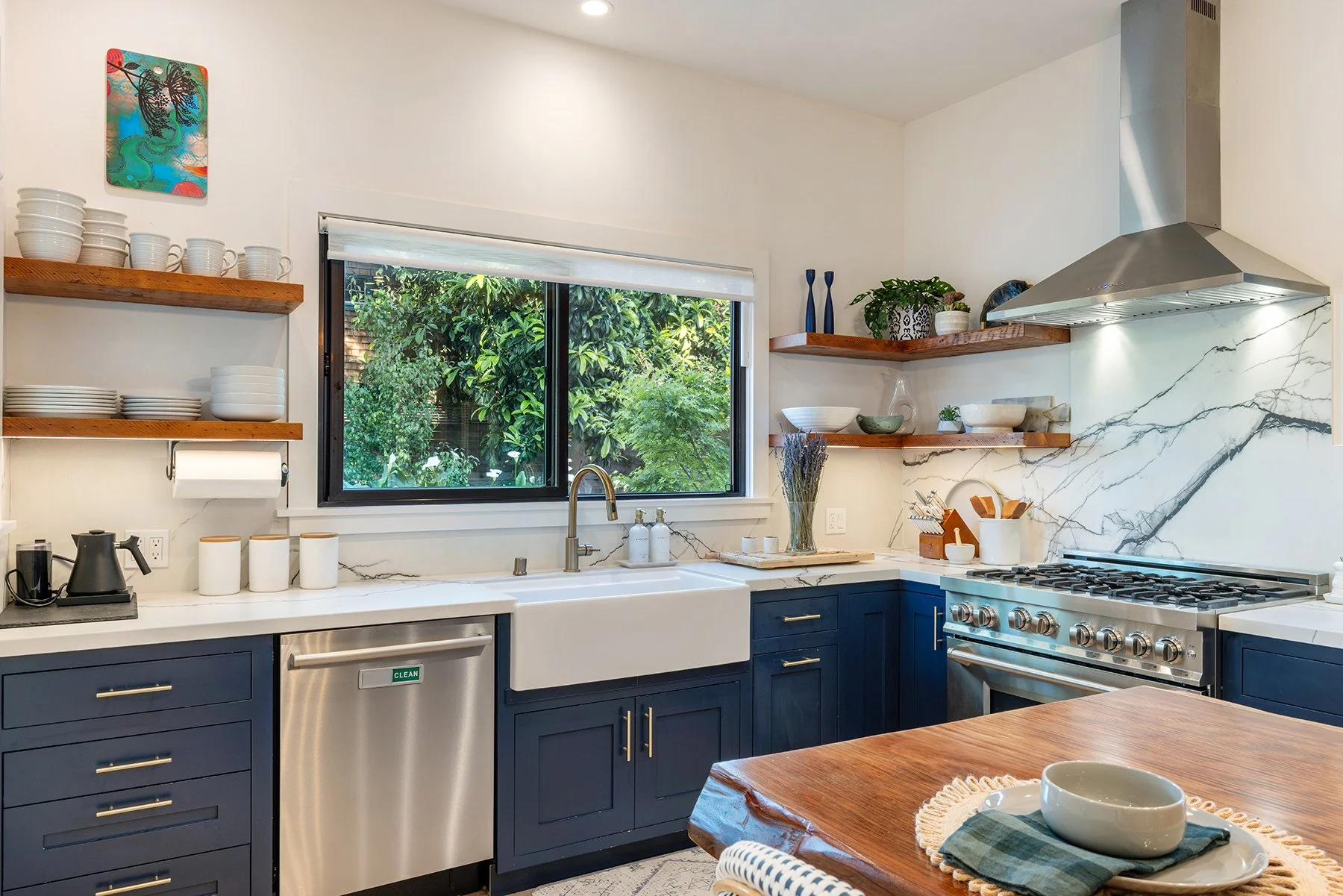 Modern kitchen with navy blue cabinets, white marble backsplash, stainless steel appliances, open wooden shelves with dishes, and a large window showing greenery outside.