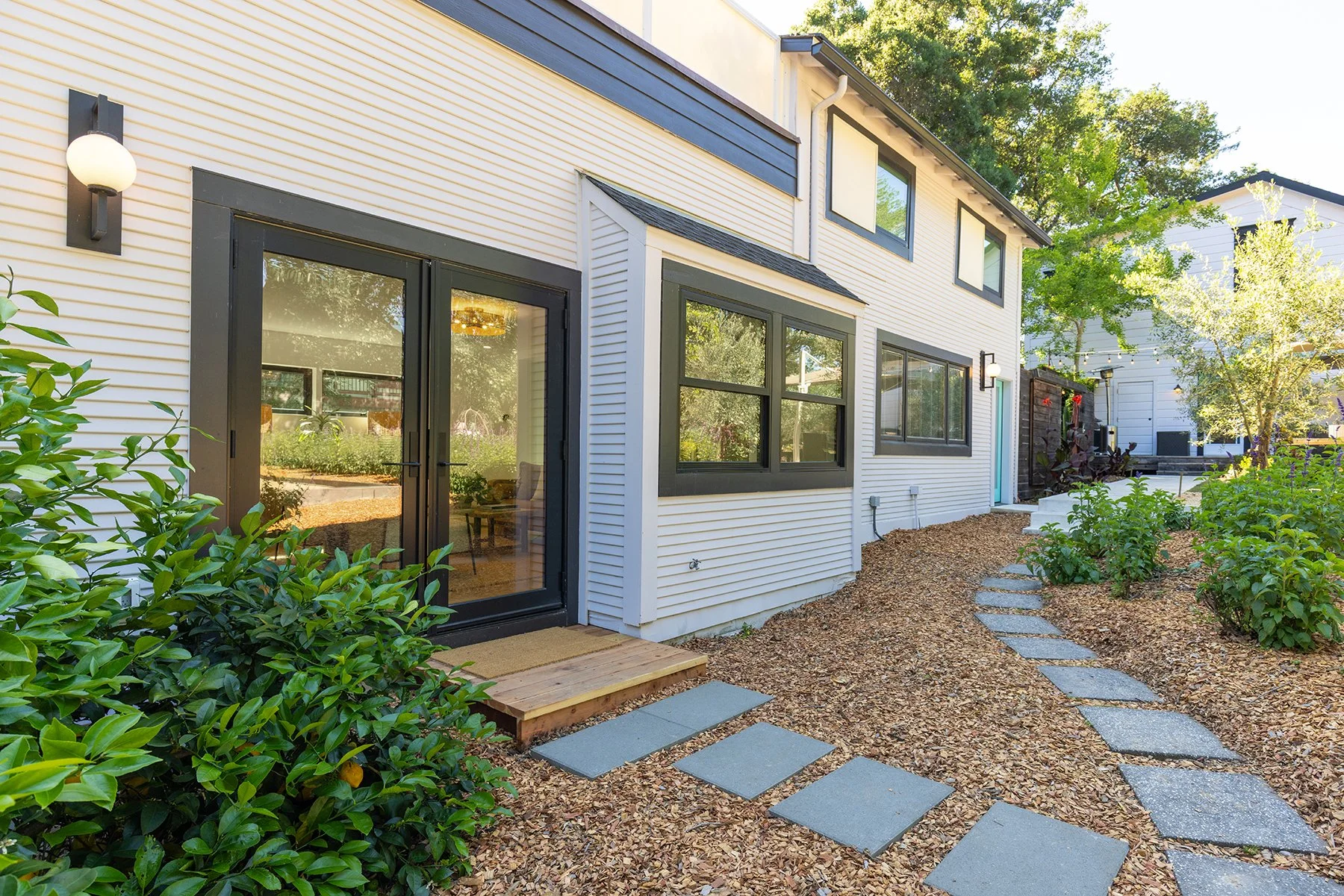 Modern two-story house with white siding, black trim, and large windows. Front yard has a stone pathway, green bushes, and trees, with outdoor lighting fixtures.