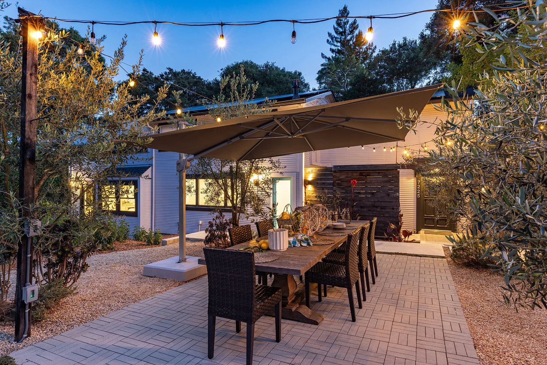 An outdoor dining area at dusk with string lights, a large umbrella over a dining table, surrounded by trees and bushes, with a house in the background.
