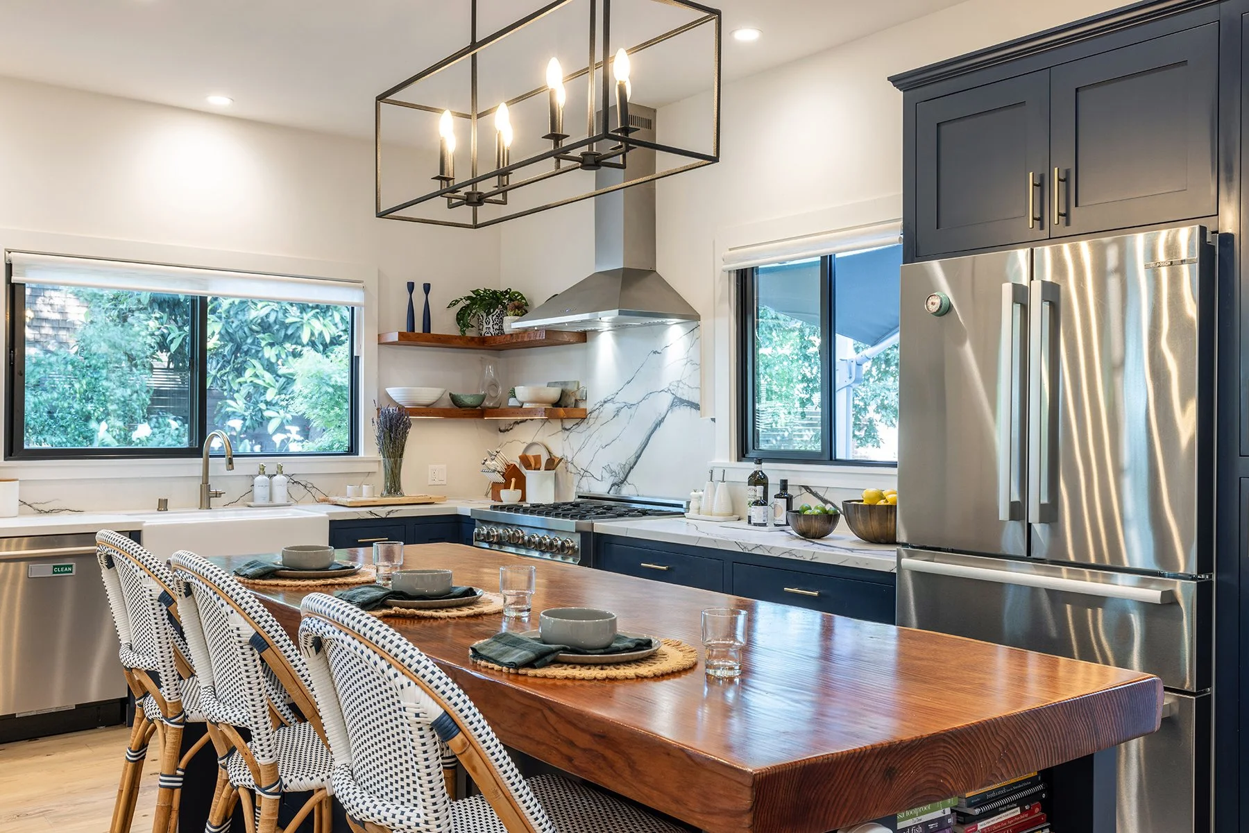 Modern kitchen with navy blue cabinets, white marble countertops, stainless steel refrigerator, open wooden shelving with bowls and decorative vases, large windows with green foliage outside, and a wooden dining table with place settings and wicker c