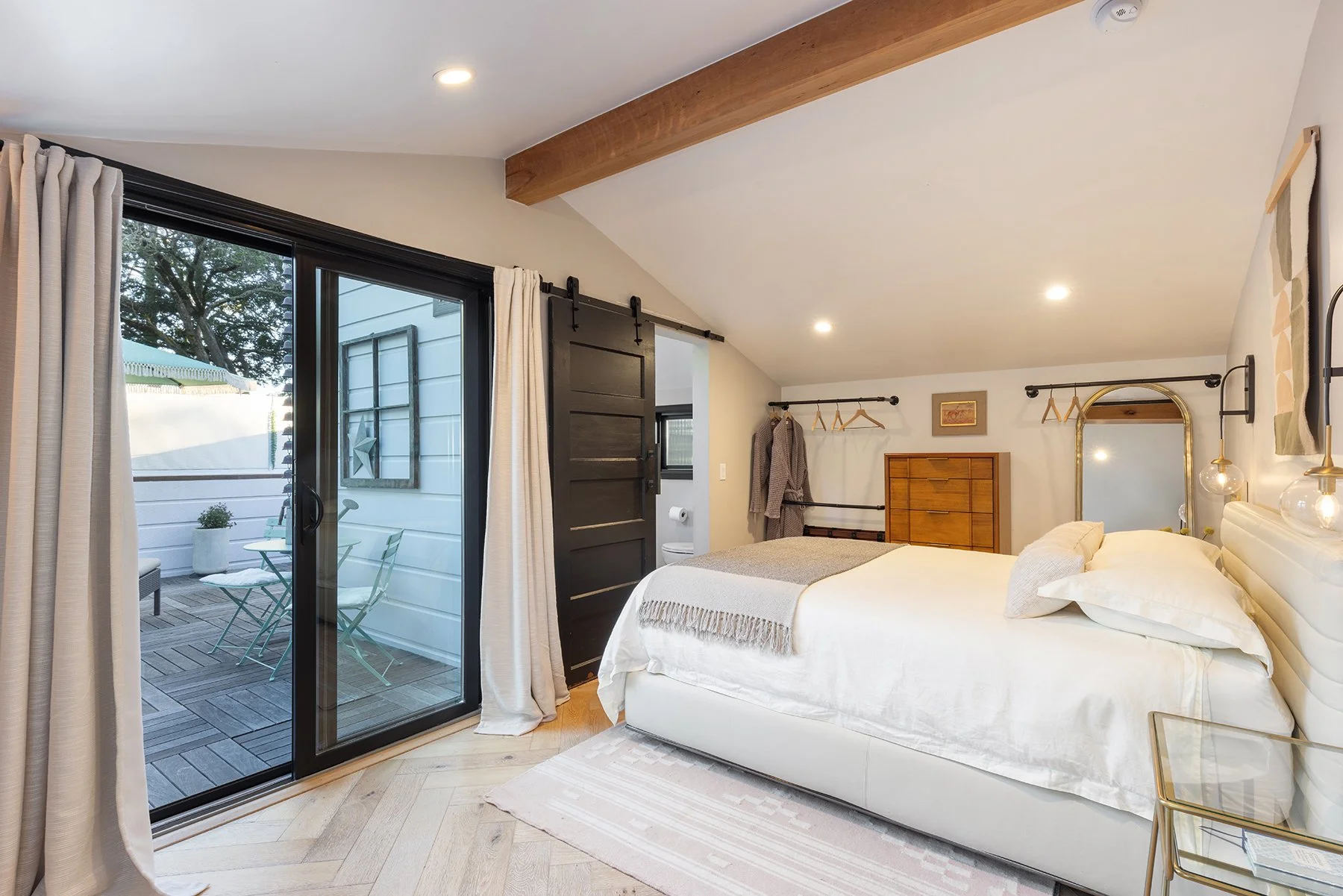 Bedroom with sliding glass door leading to a patio, with an outdoor table and chairs. The bedroom has a white bed with a beige blanket, a wooden dresser, and wall-mounted hangers. The room features wooden elements, including a ceiling beam and hardwo