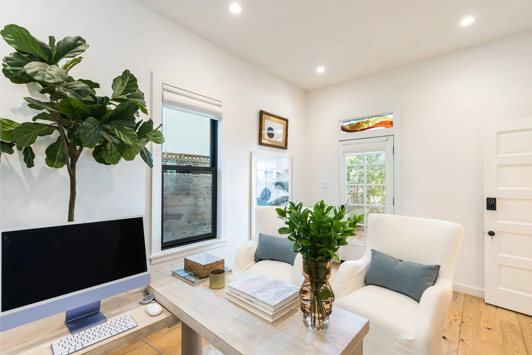 A bright living room with white walls, large windows, a desk with a computer, a plant on the coffee table, white chairs with gray pillows, framed artwork, and a door leading outside.