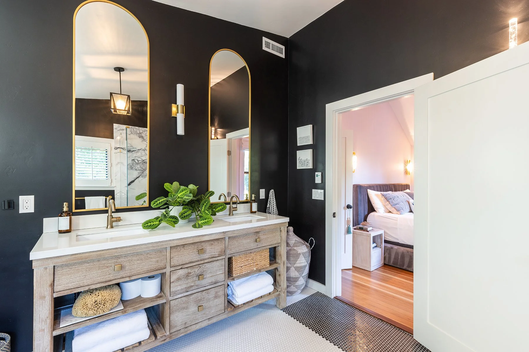 Modern bathroom with a wooden vanity, two large mirrors with gold trim, black walls, a sink with a gold faucet, a green plant on the counter, and a view into a bedroom with pink walls and a bed.