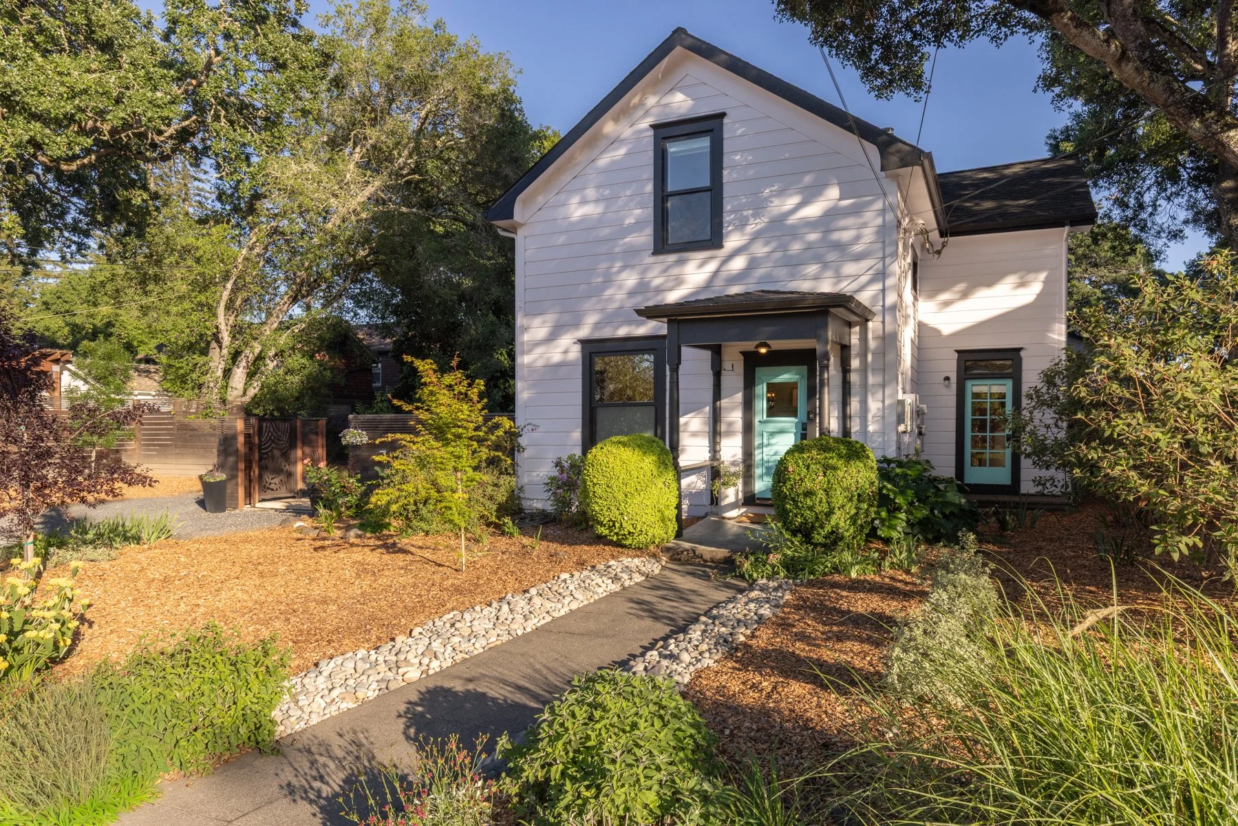 White two-story house with black trim and a light blue door, surrounded by a lush garden with various plants and trees, a pathway lined with white stones leading to the front door, and a wooden fence gate on the left.