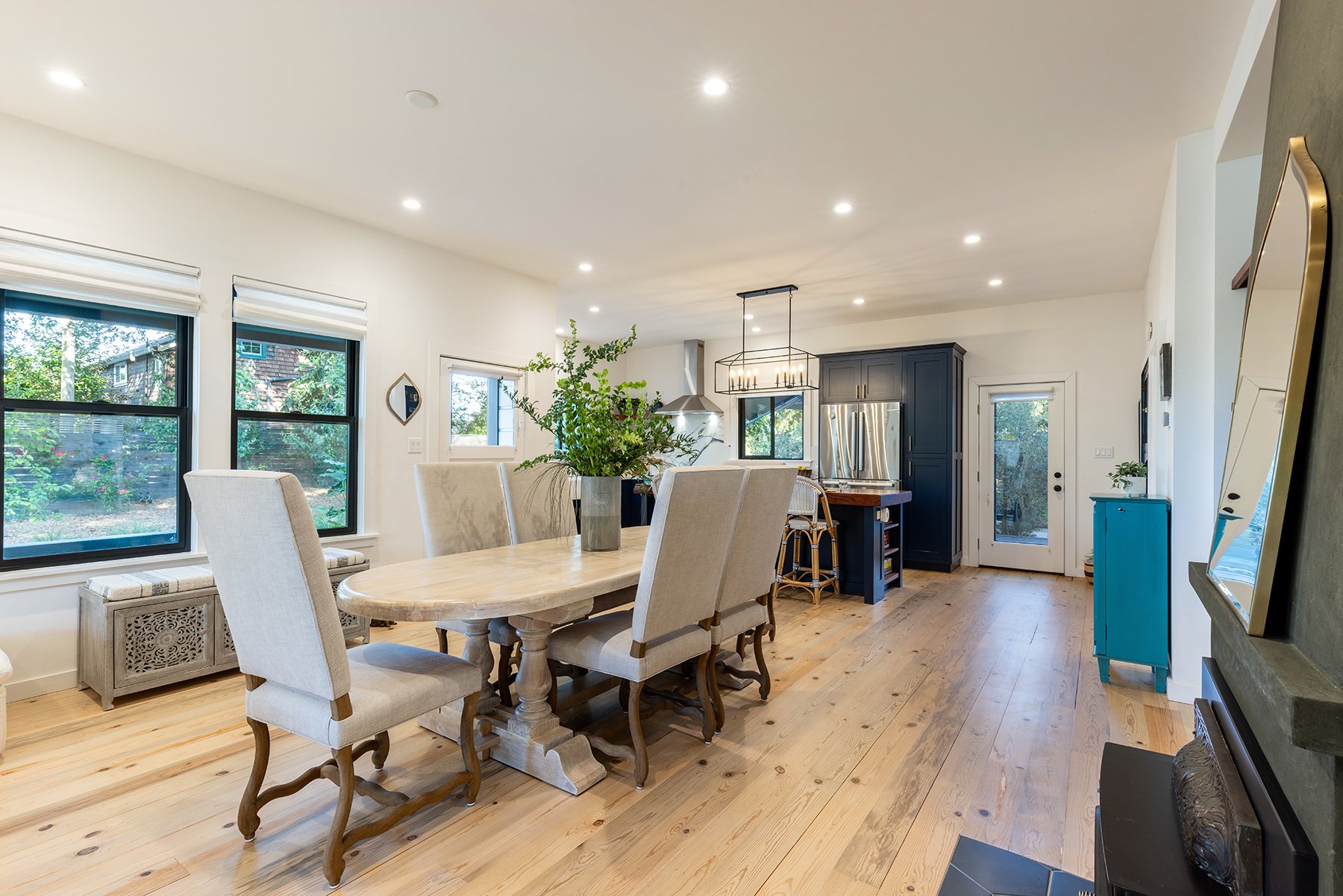 Open-concept kitchen and dining area with large windows, a dining table with beige chairs, a blue cabinet, and modern appliances.