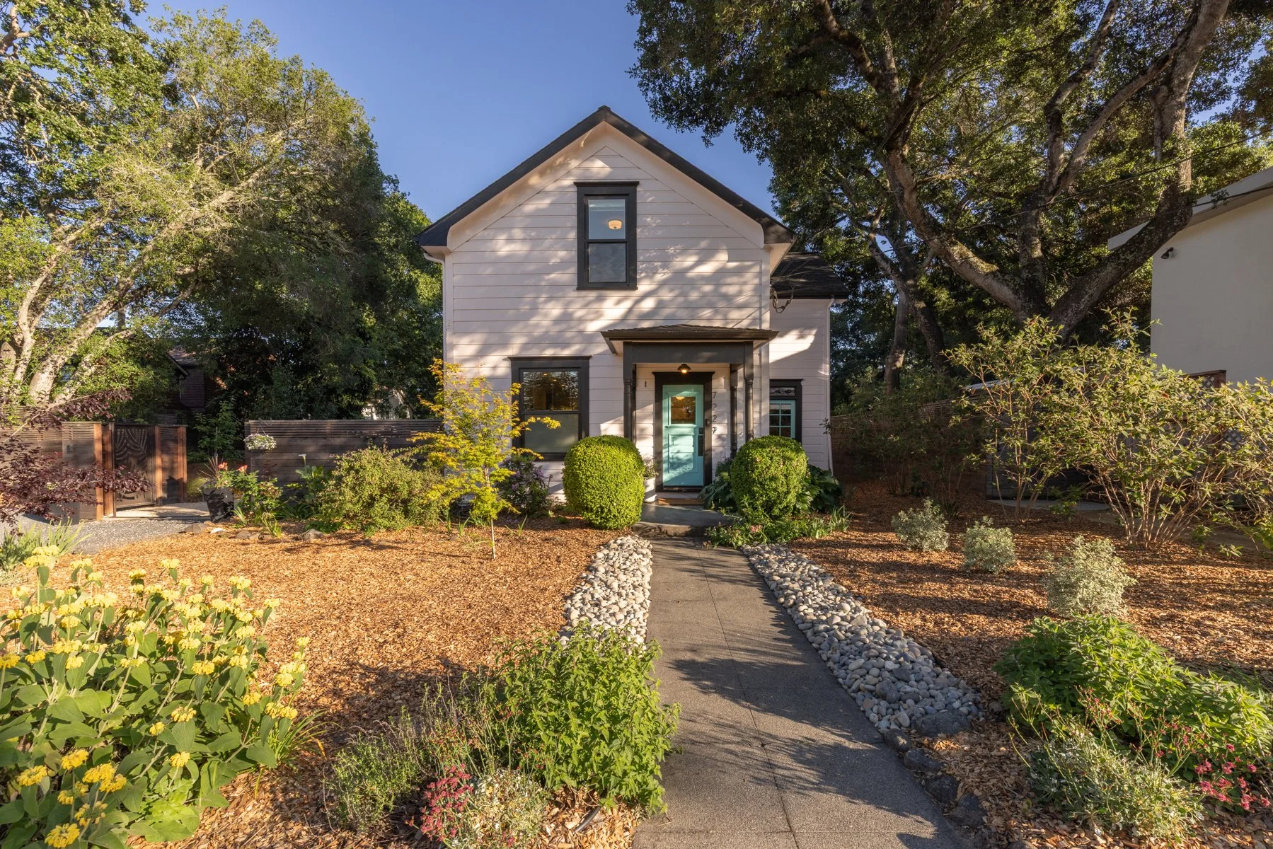 A cozy two-story white house with black window frames and a light blue front door, surrounded by a landscaped garden with shrubs, trees, and a pathway lined with white rocks under a clear blue sky.