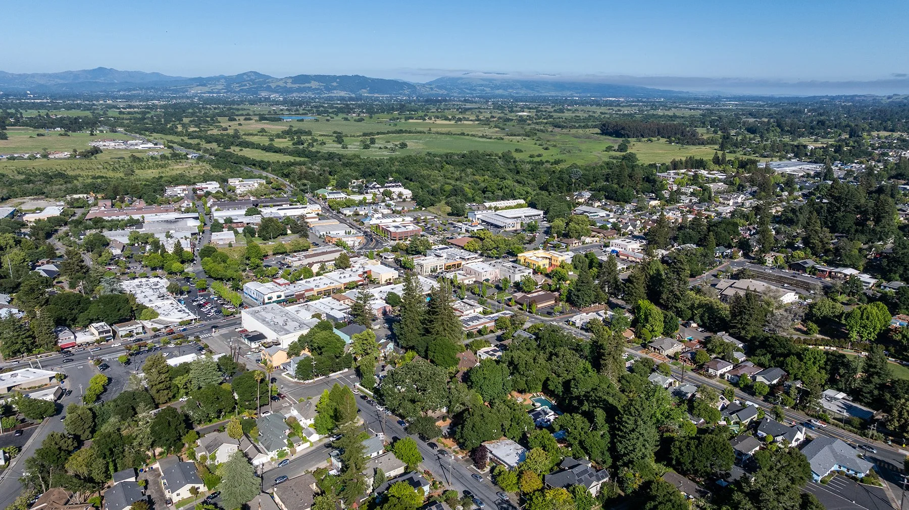 Aerial view of a small town with commercial and residential buildings, surrounded by green trees, open fields, and mountains in the distance under a clear blue sky.