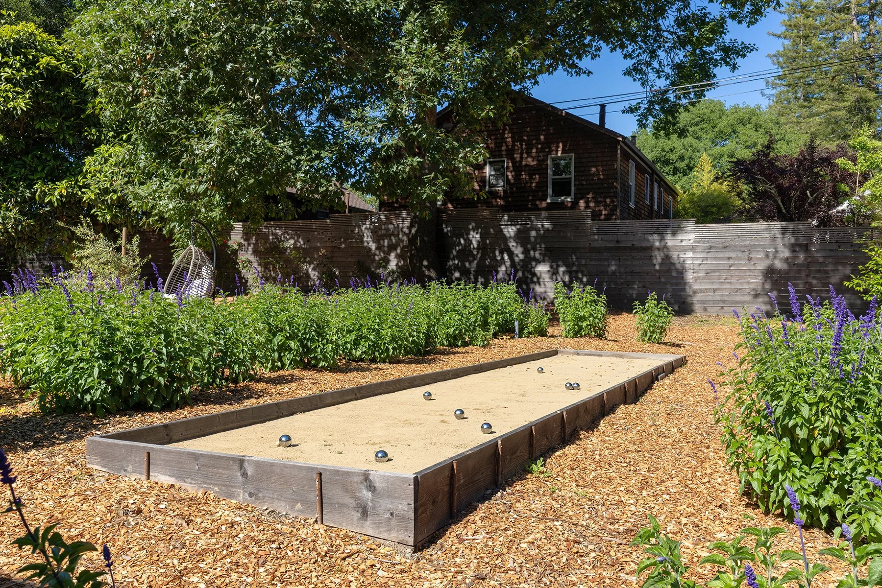 A backyard with a sand bocce court containing metallic balls, surrounded by purple flowers and green bushes, with a large tree providing shade and a wooden fence and house in the background.