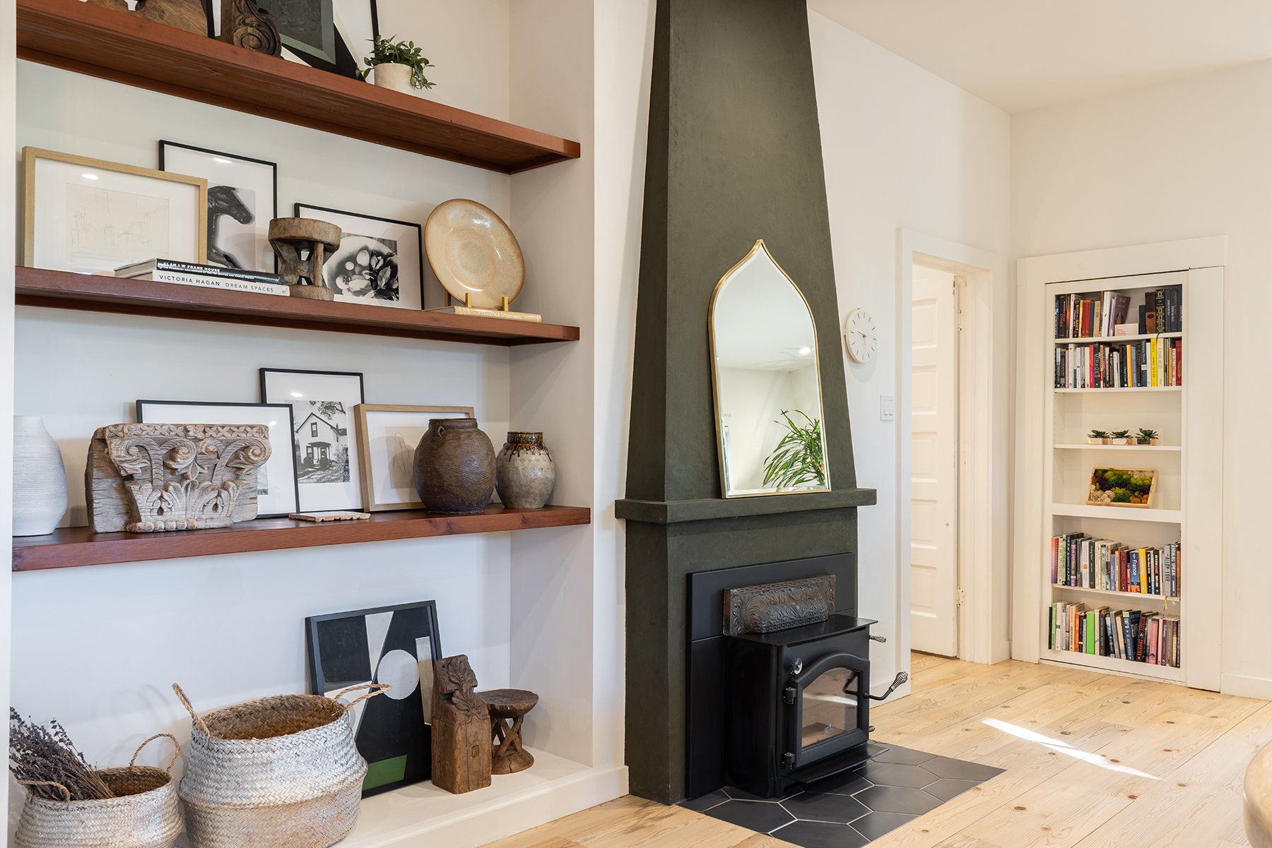 Living room interior with two wooden shelves holding framed art, pottery, and decorative items, a mirror on a dark gray fireplace mantle, and a white bookshelf filled with books and small plants.