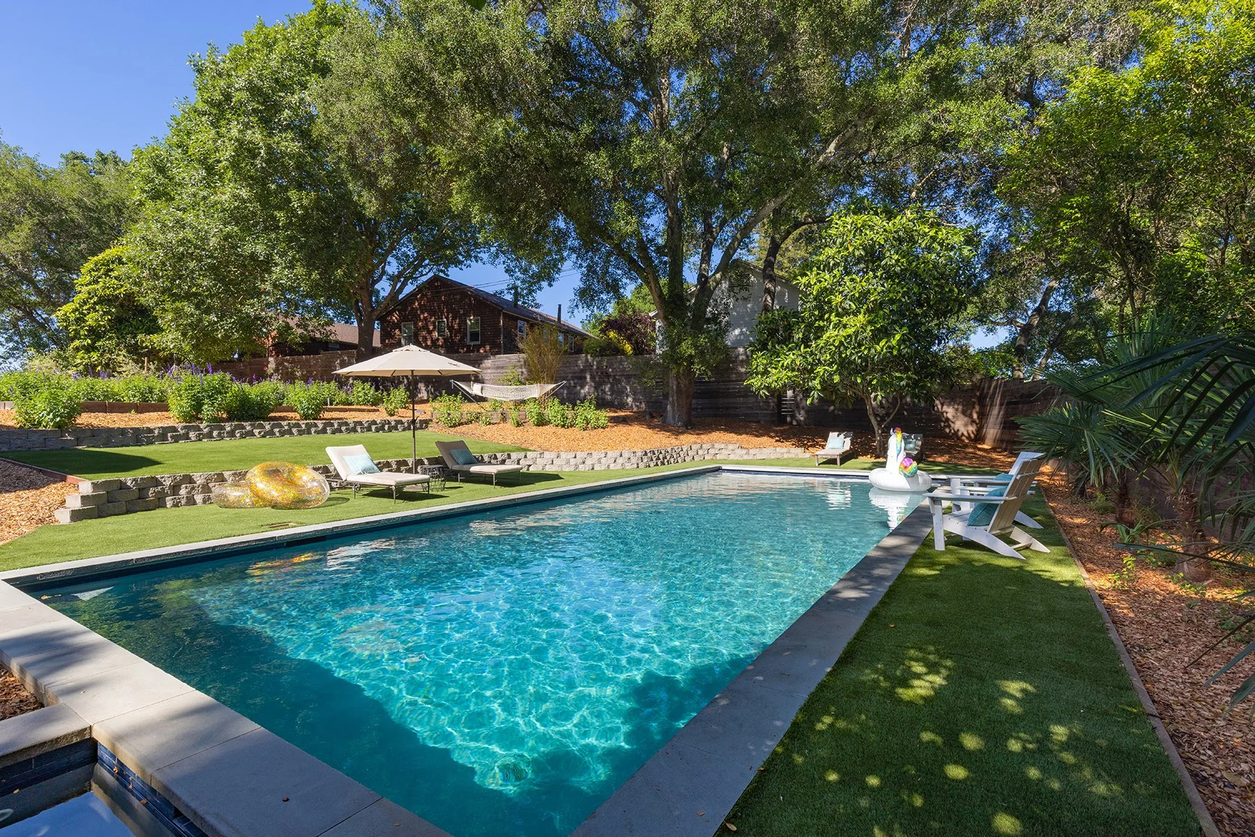 Backyard swimming pool scene with lounge chairs, a large tree providing shade, a patio umbrella, inflatable swan and donut floaties, and lush greenery surrounding a residential house.
