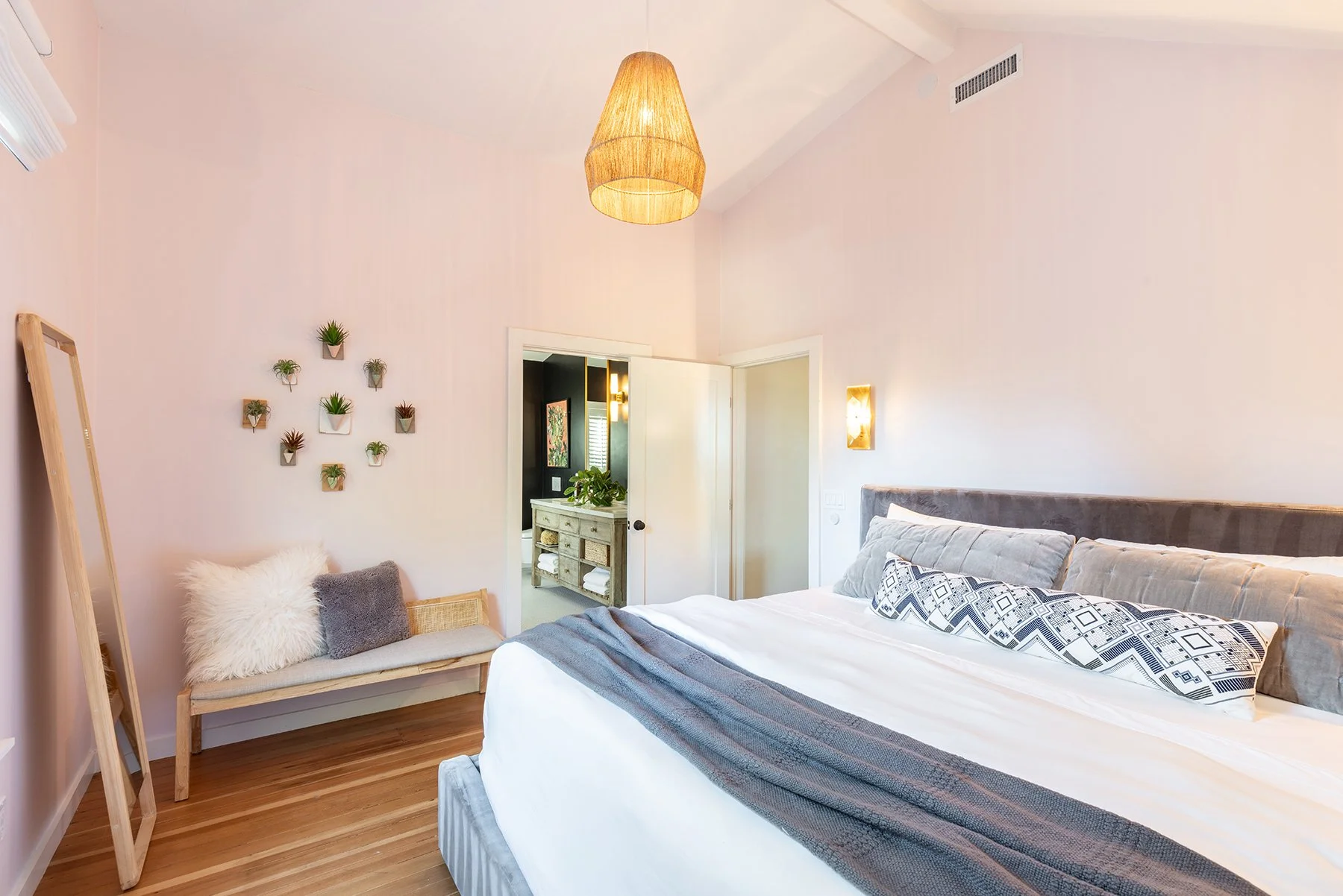 A bedroom with a bed dressed in white linens and decorative pillows, a bench with fuzzy throw pillows, wall decor with small potted plants, a wooden mirror leaning against the wall, and a ceiling light fixture.