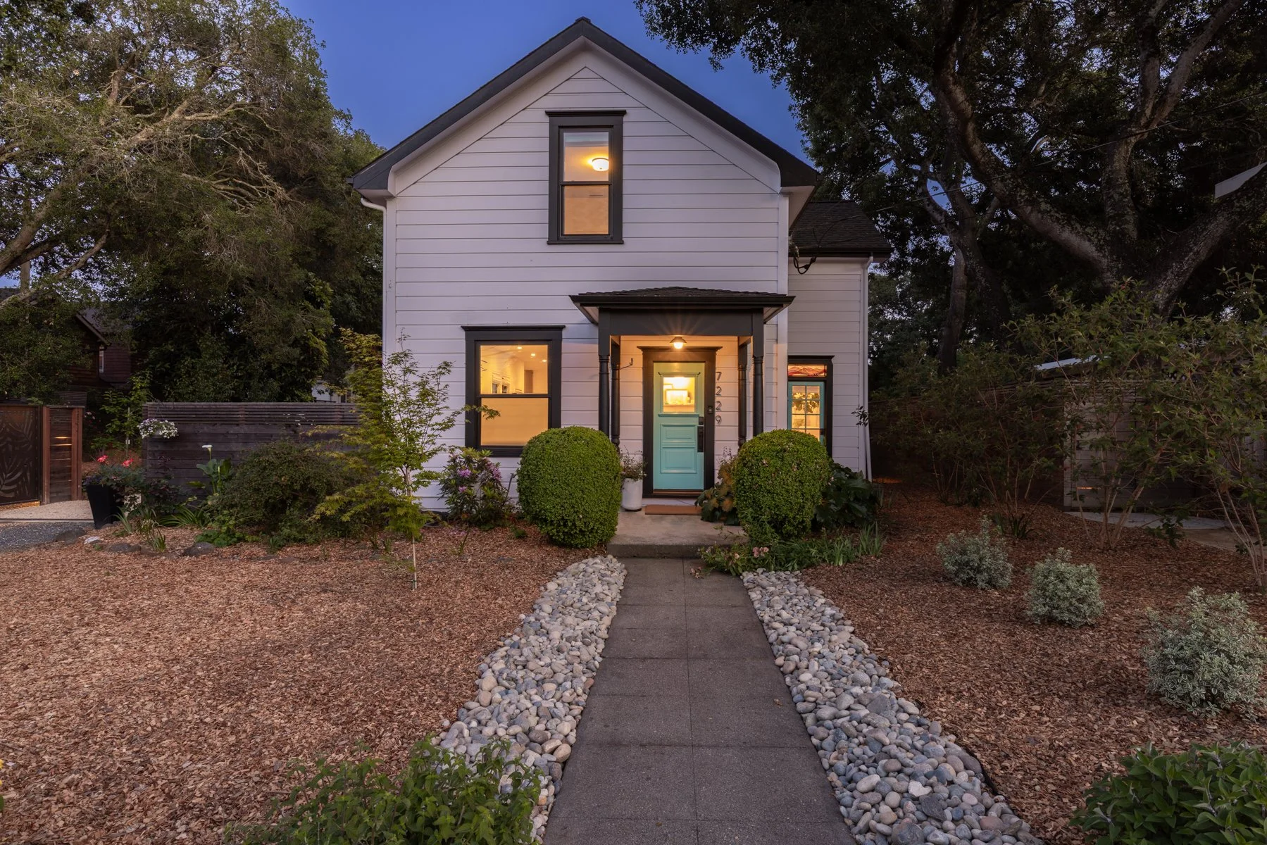 Front view of a white two-story house with black trim and a light blue front door, surrounded by bushes and small plants, with a gravel walkway leading to the entrance, evening lighting, and trees in the background.