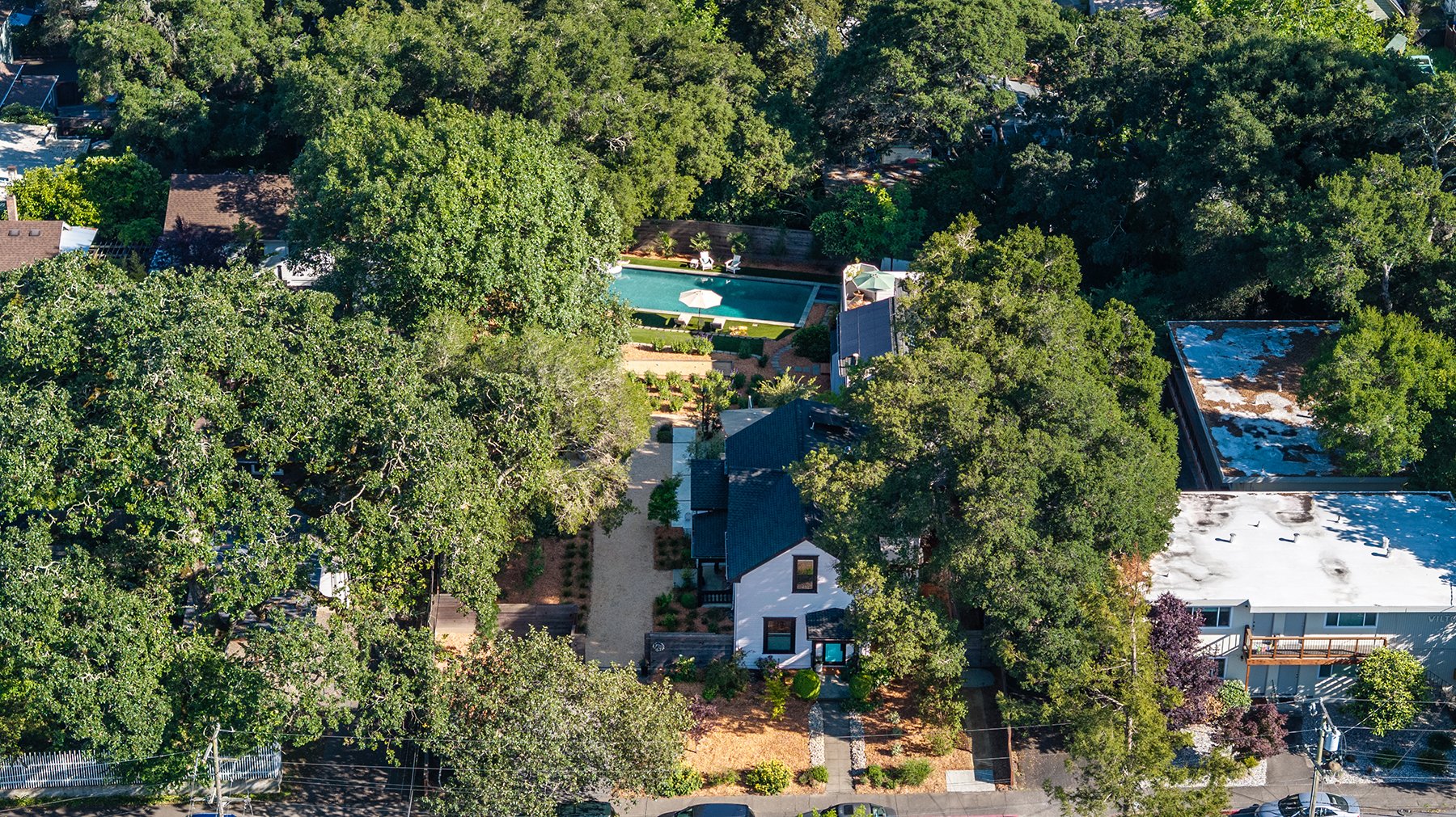 Aerial view of a residential neighborhood with houses, trees, and a central backyard pool area with lounge chairs and a white umbrella.