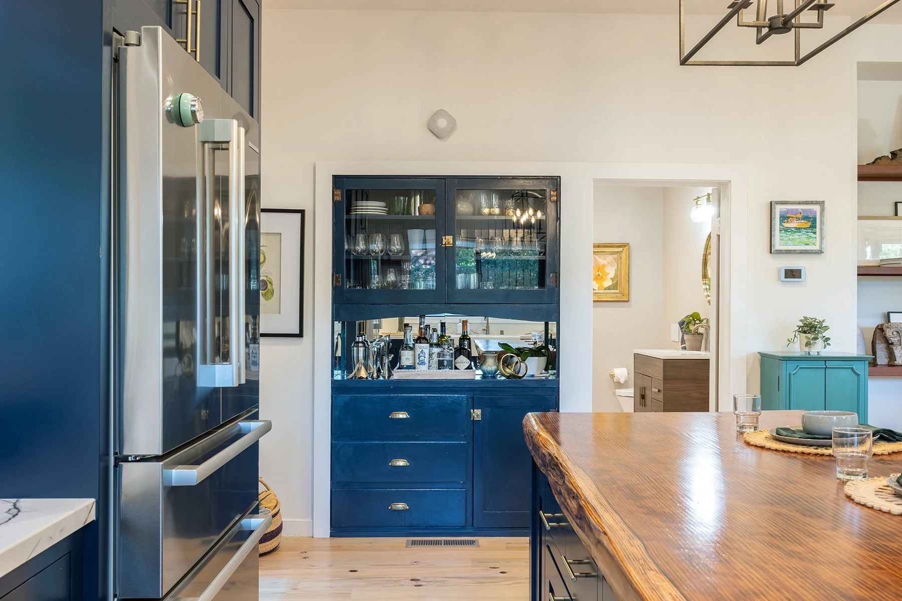 A kitchen scene with a blue cabinet filled with glassware and bottles, a wooden dining table with placemats and dishes, and artwork on the walls.