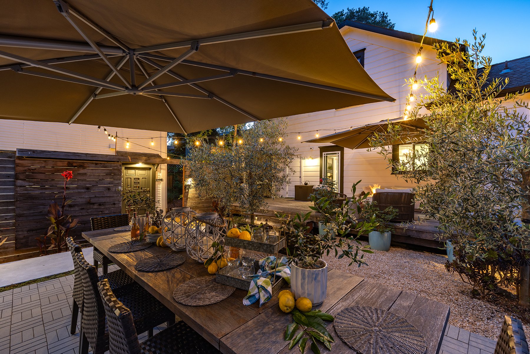 A cozy outdoor patio scene during twilight with string lights, large umbrellas, a dining table set with woven placemats, glassware, and decorative items, surrounded by potted plants, trees, and a wooden deck with a house in the background.