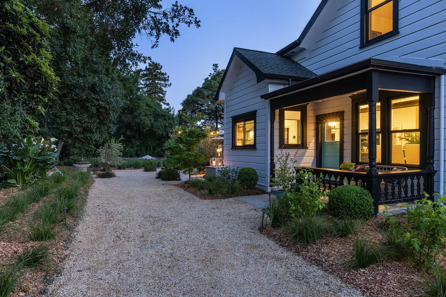 A cozy white house with black trim and a small porch, illuminated from within, surrounded by a lush garden and a gravel pathway during dusk.