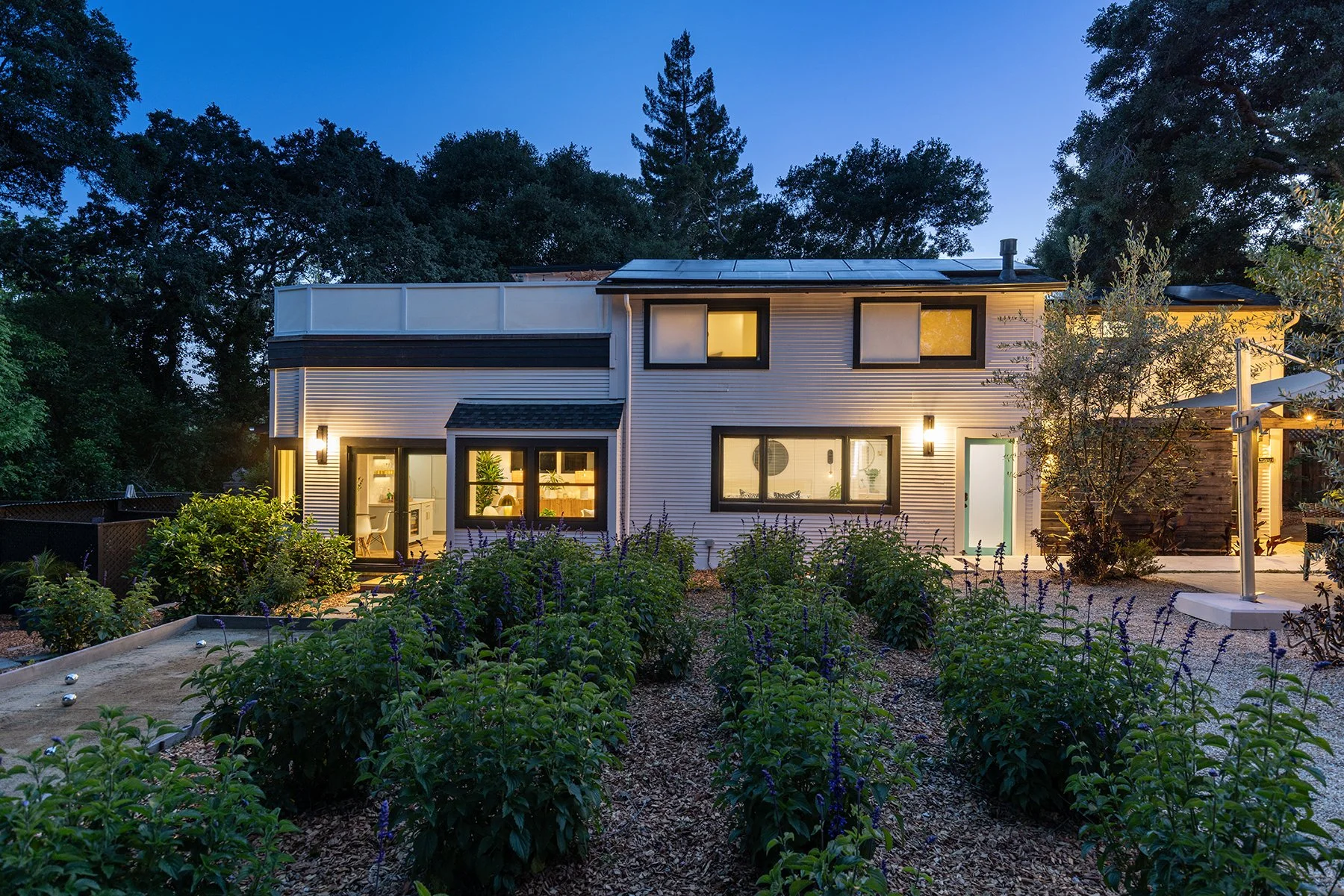 A modern two-story house with illuminated windows at dusk, surrounded by greenery and a garden with blooming plants. The house has solar panels on the roof and outdoor lighting.