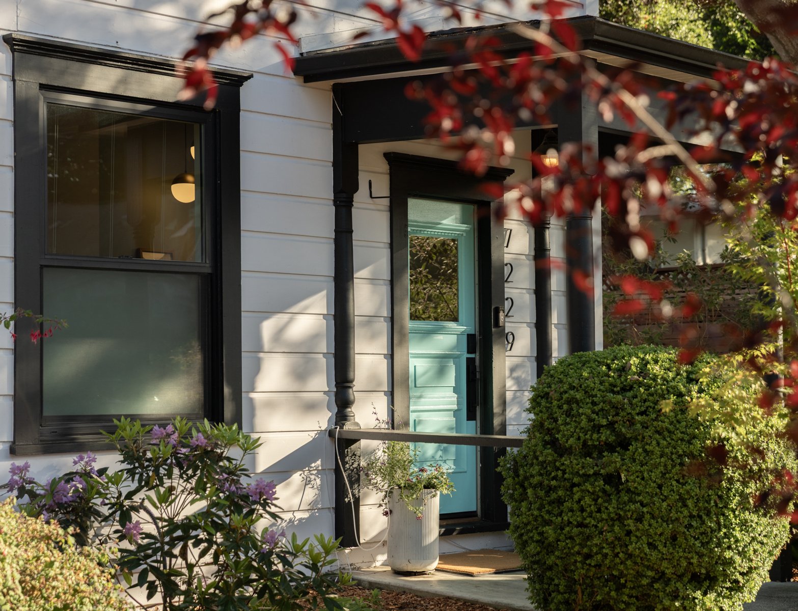 Close-up of a house entrance with a turquoise door, black trim, and black front porch columns, surrounded by lush green shrubs and plants.