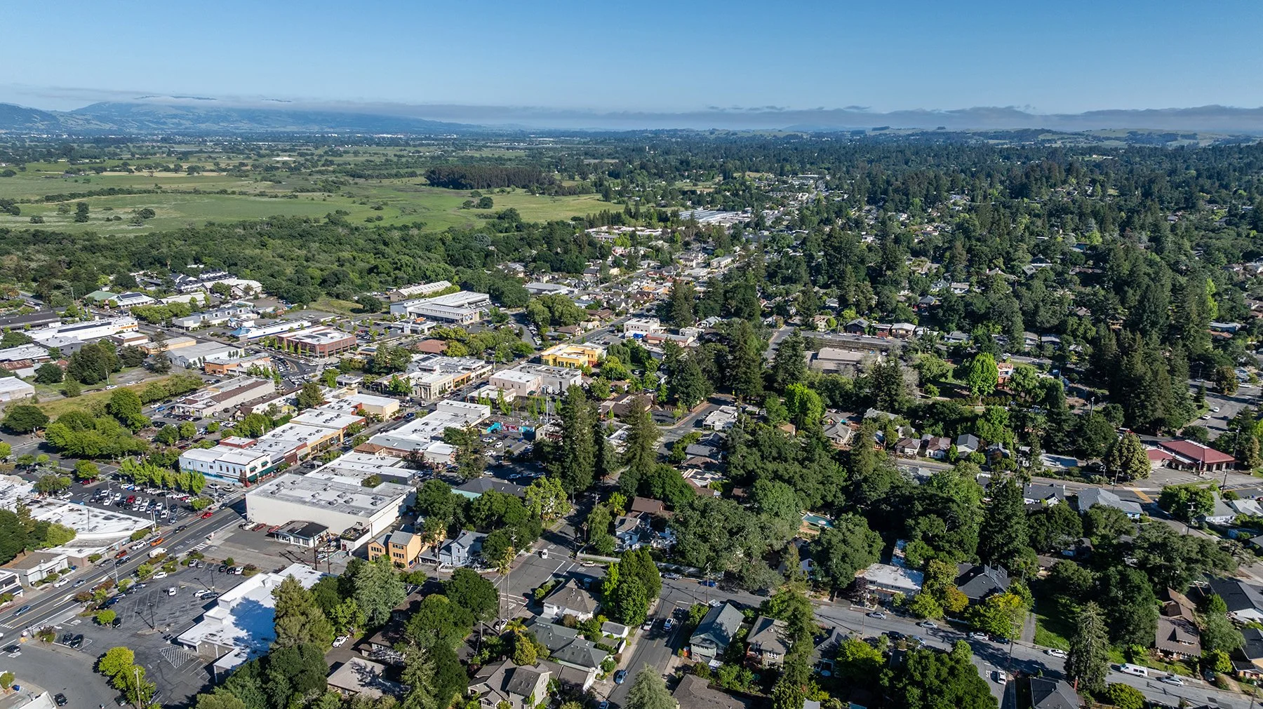 An aerial view of a small town surrounded by green trees and open fields, with a mountain range in the background under a clear blue sky.