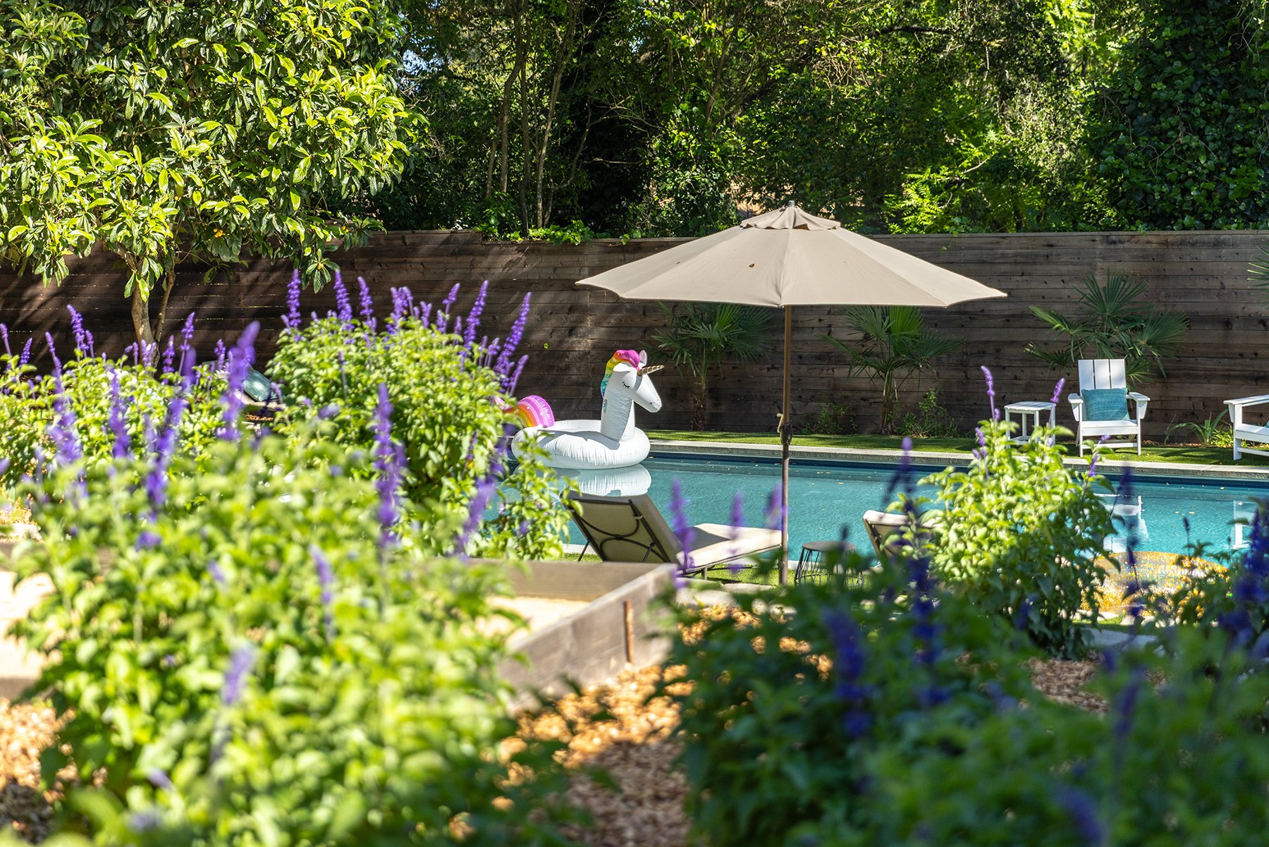 A backyard swimming pool area with a float shaped like a unicorn, surrounded by lush green plants, purple flowers, patio furniture, and a large beige umbrella.