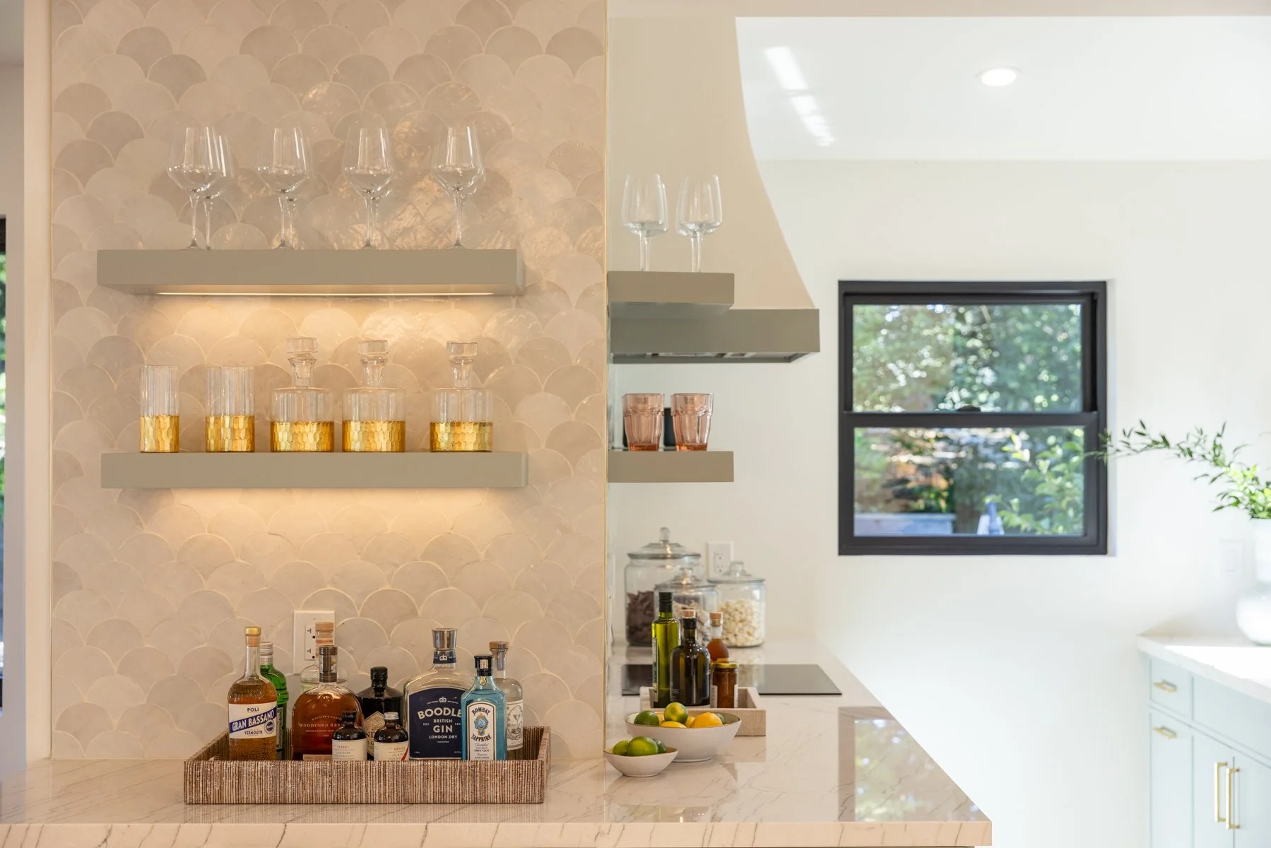A kitchen counter with a basket of assorted liquor bottles, bowls with lemons and limes, and jars of popcorn and dried goods. Behind the counter, beige shelves with wine glasses and tumblers, a window showing greenery, and a white wall with a small p