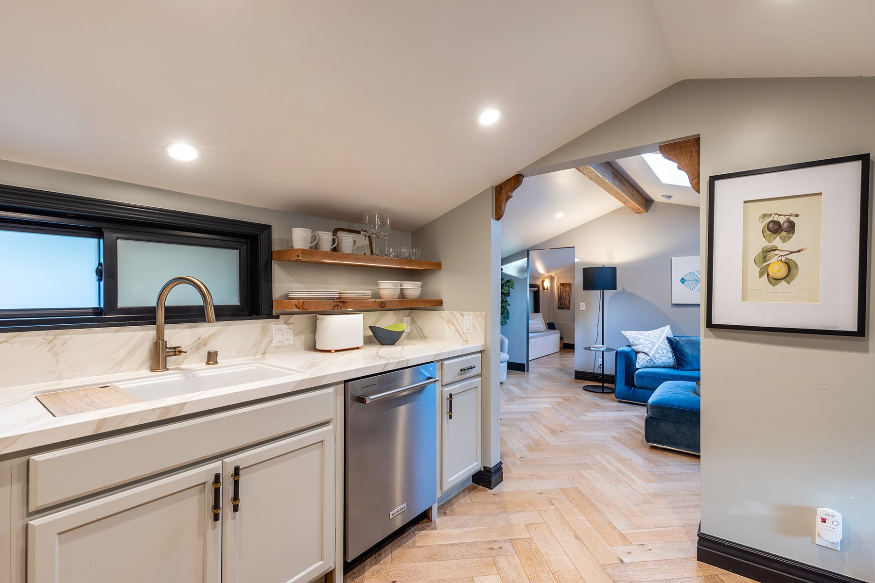 Modern kitchen with white cabinets, marble countertops, open wooden shelves with dishes, and a view into a cozy living space with a blue sofa, artwork, and a floor lamp.