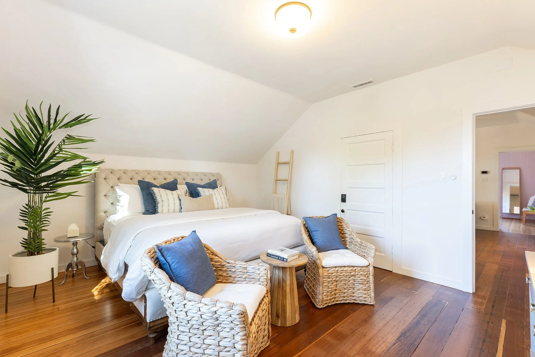 Bright bedroom with white walls, wood flooring, and a large bed with a tufted headboard decorated with blue and beige pillows. To the left, there's a tall green plant in a white pot and a small round side table. Wicker chairs with blue pillows are po