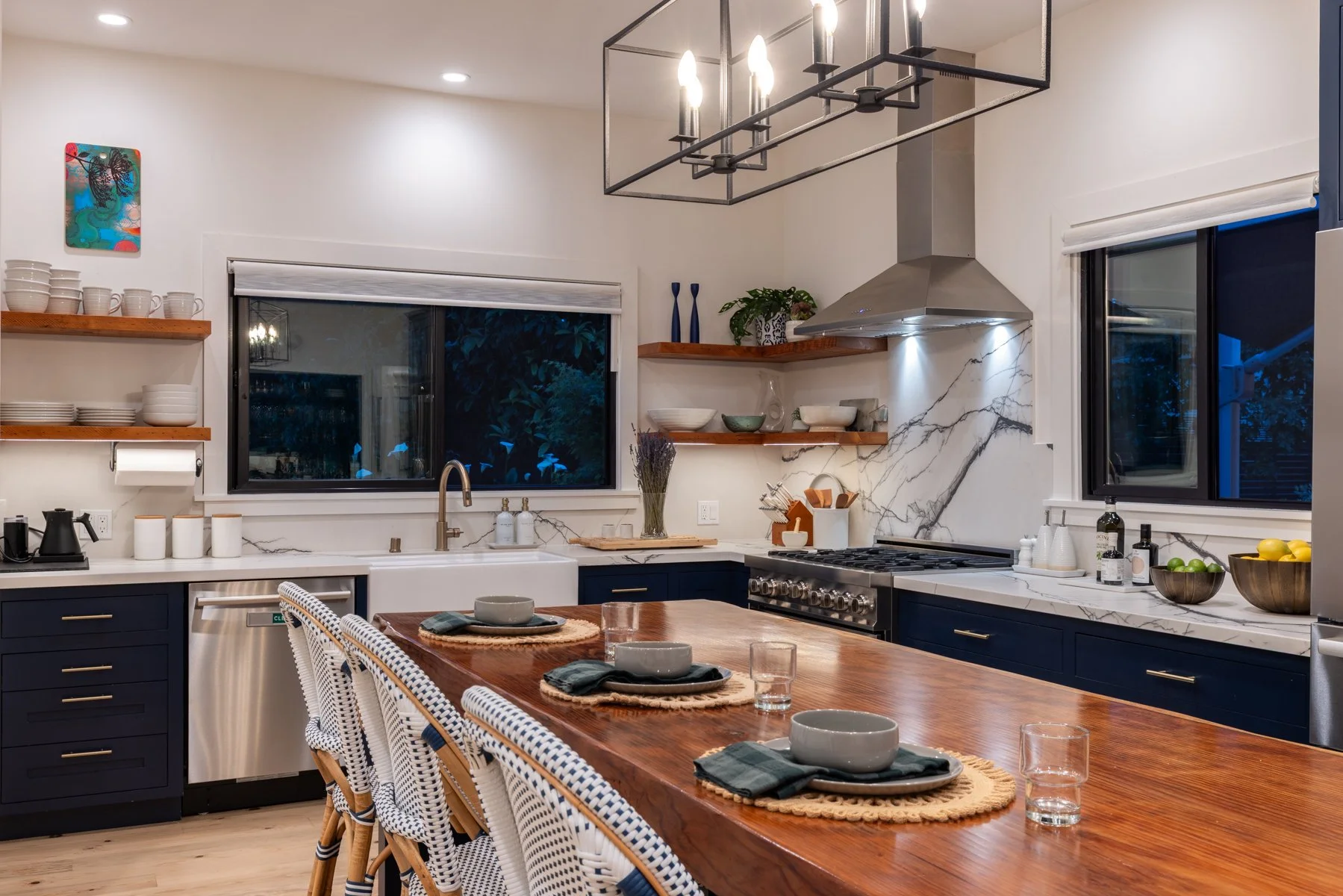 Modern kitchen with wooden table, navy blue cabinets, marble backsplash, open shelves with dishes and decor, stainless steel range, and large windows.
