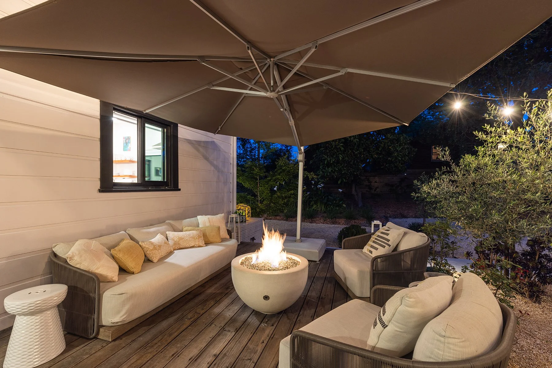 An outdoor patio at night with a beige sofa, armchairs, and throw pillows, a fire pit, a large tan umbrella, and garden lighting.