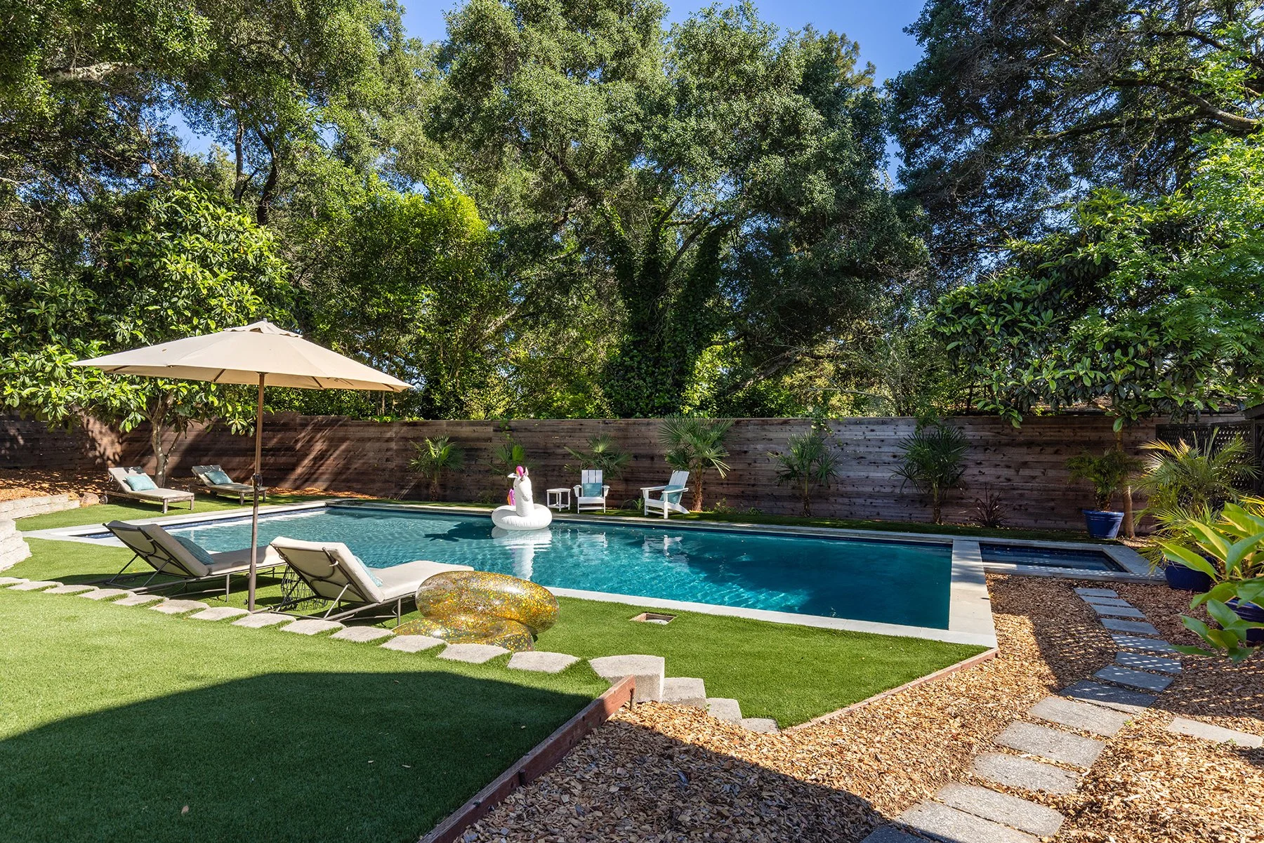 A backyard with a swimming pool surrounded by green grass and plants, with lounge chairs, a large umbrella, and an inflatable unicorn floating in the pool, and tall trees in the background.