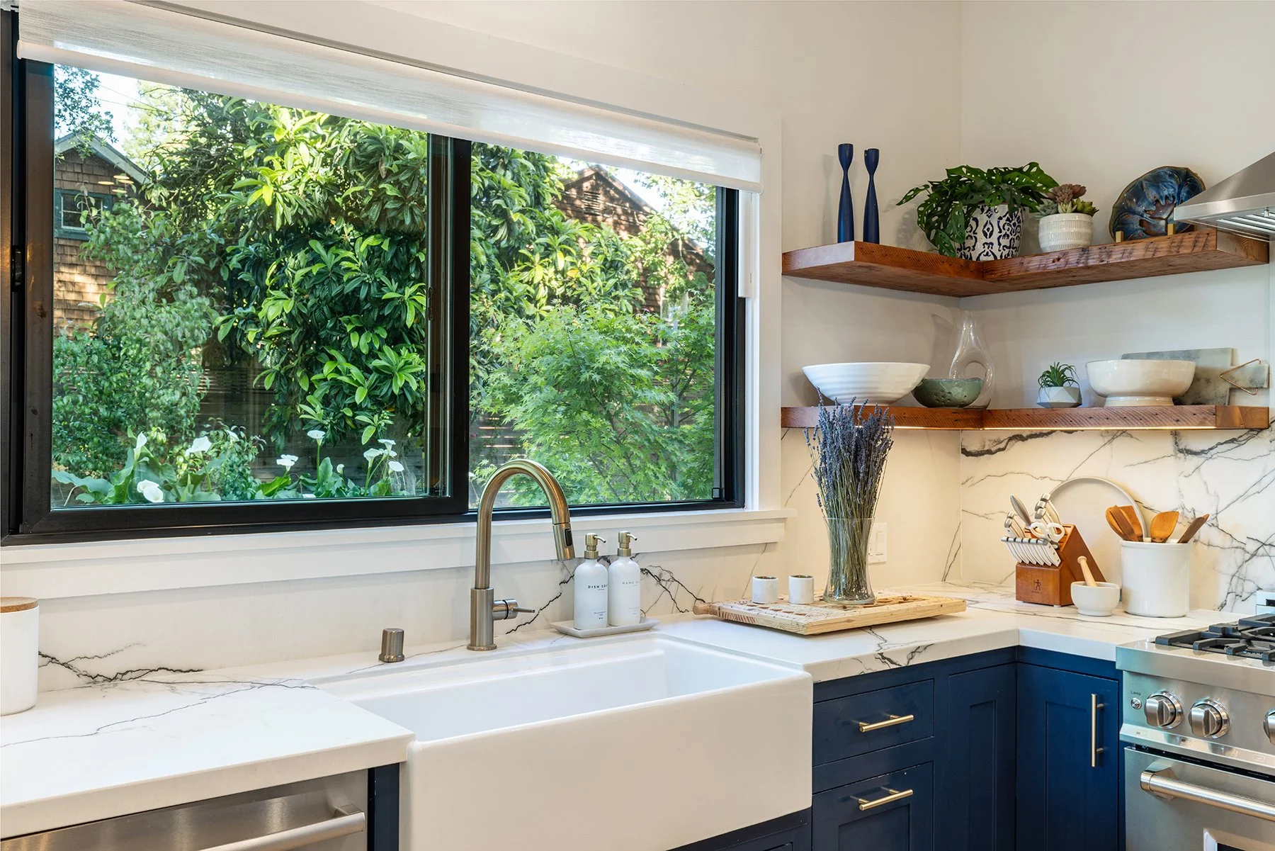 Kitchen with a large window showing green trees outside, white marble countertops, a white farmhouse sink, brass faucet, and open wooden shelves with decorative dishes, plants, and vases.
