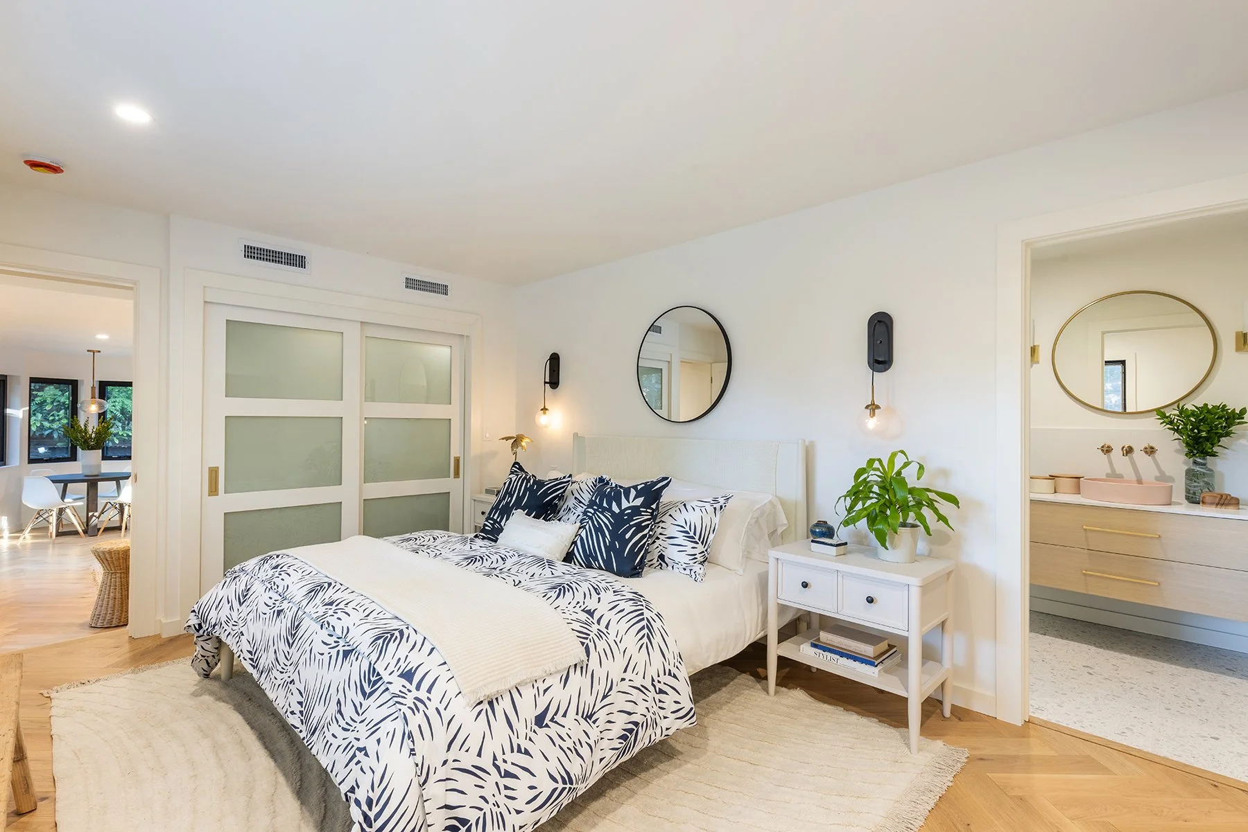 A modern bedroom with a white bed adorned with navy and white botanical-patterned pillows, a white nightstand with books and a potted plant, a round mirror above the bed, and a bathroom with a pink sink and a large mirror visible through an open door