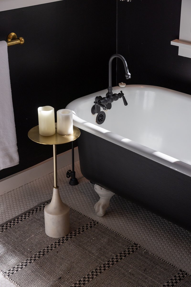 A bathroom with a black clawfoot bathtub, a black faucet, a small side table with two candles, a white towel hanging on a gold towel rack, and patterned floor tiles.