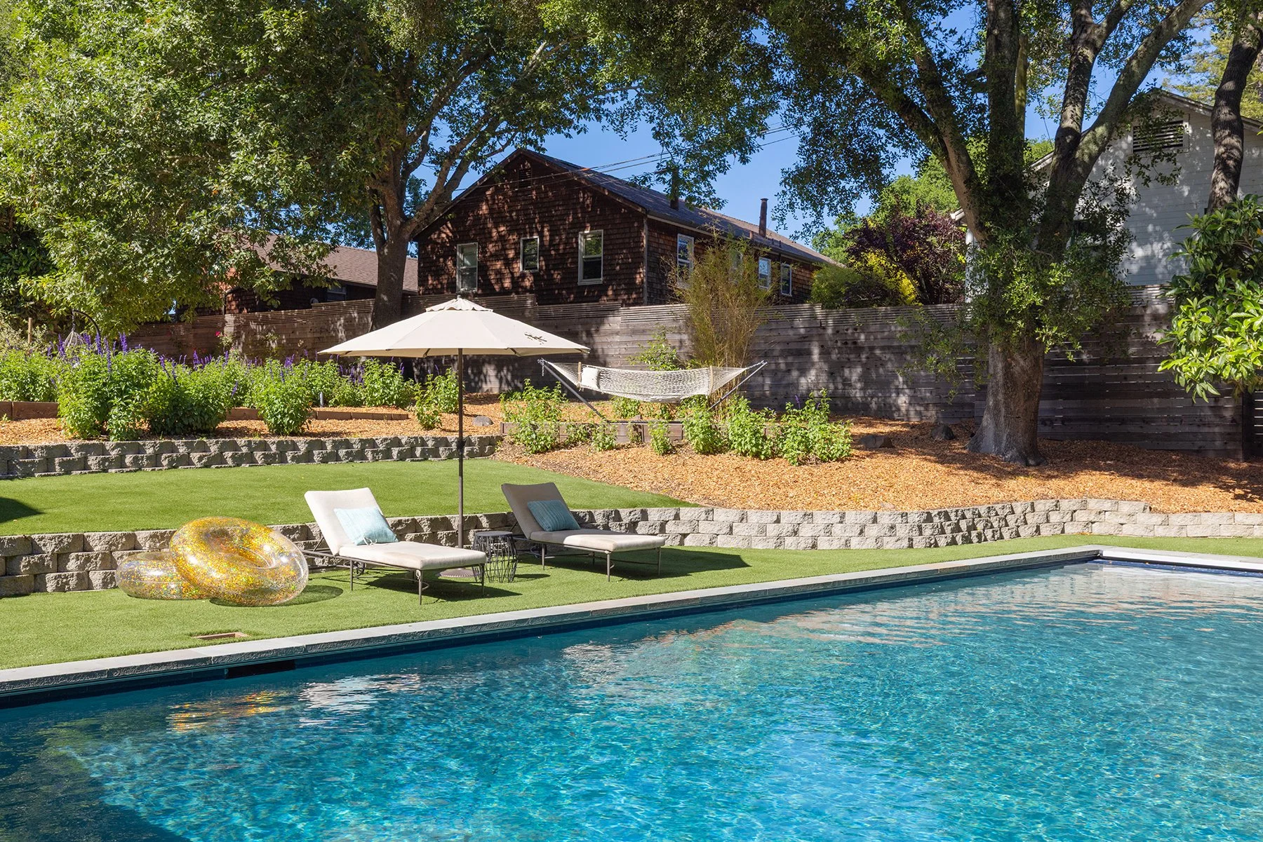 Backyard with swimming pool, lounge chairs, umbrella, and garden area with trees and bushes.