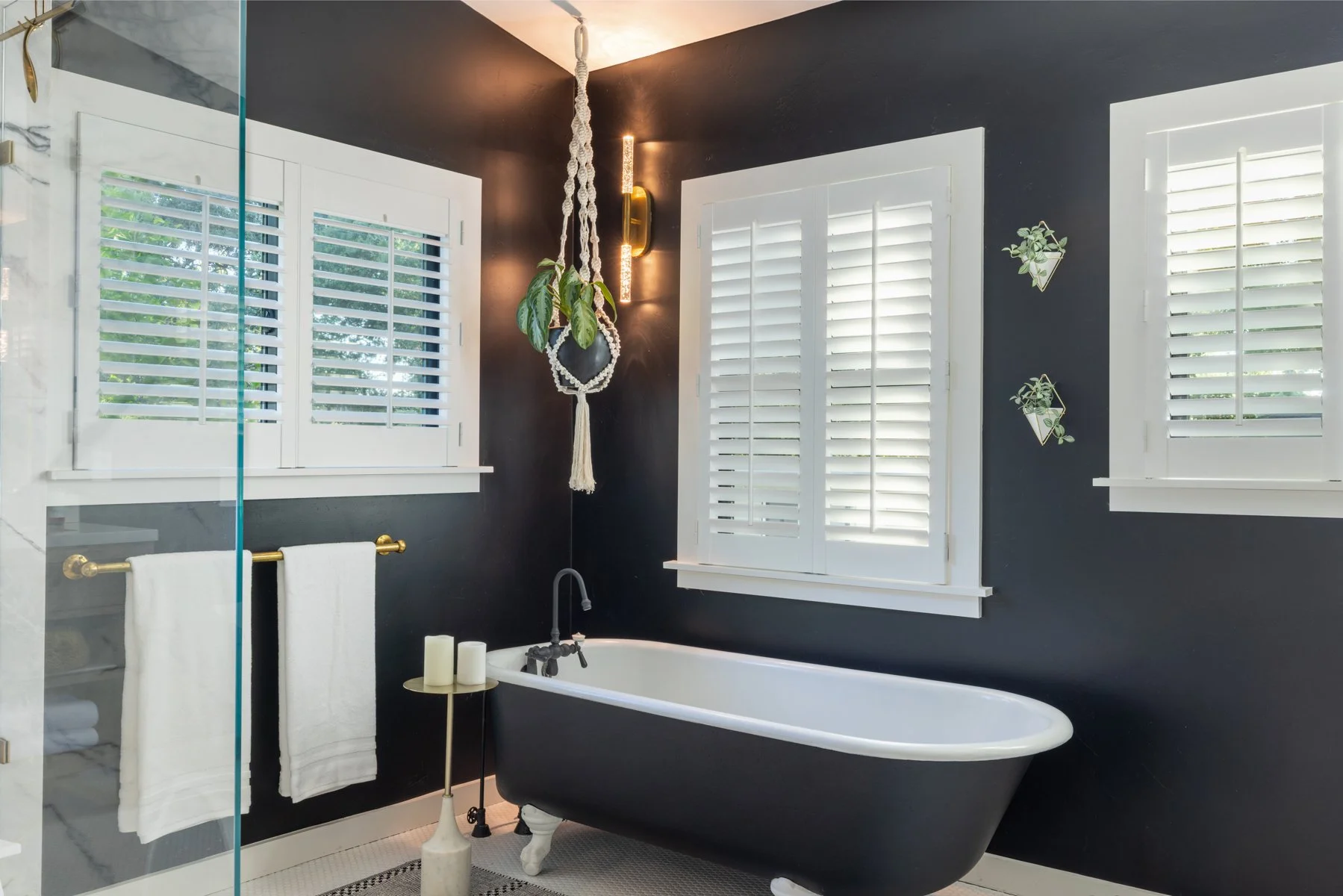 A black clawfoot bathtub in a bathroom with dark blue walls, white window shutters, and a gold towel rack with white towels. A round side table with candles, a hanging potted plant, and a modern wall light next to the window.