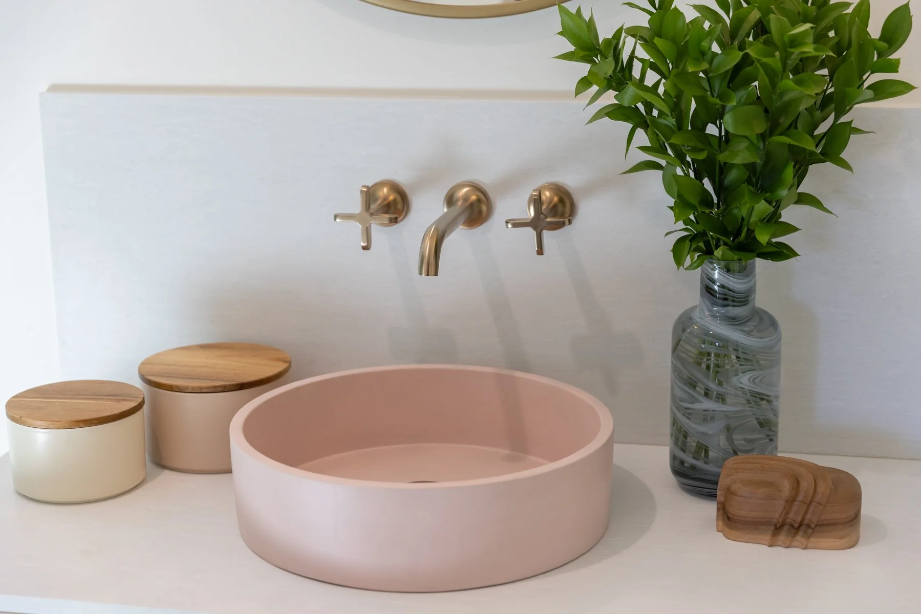 A bathroom sink area with a pink basin, a tall marbled vase with green leafy branches, and two small wooden and cream-colored containers with wooden lids.