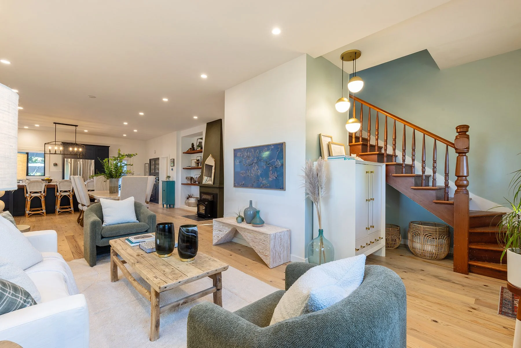 Living room with white and green sofas, wooden coffee table with black and gold decorative vases, a small marble-top table with vases, a wooden dining table with wicker chairs, a kitchen with black cabinets and stainless steel appliances, and a stair