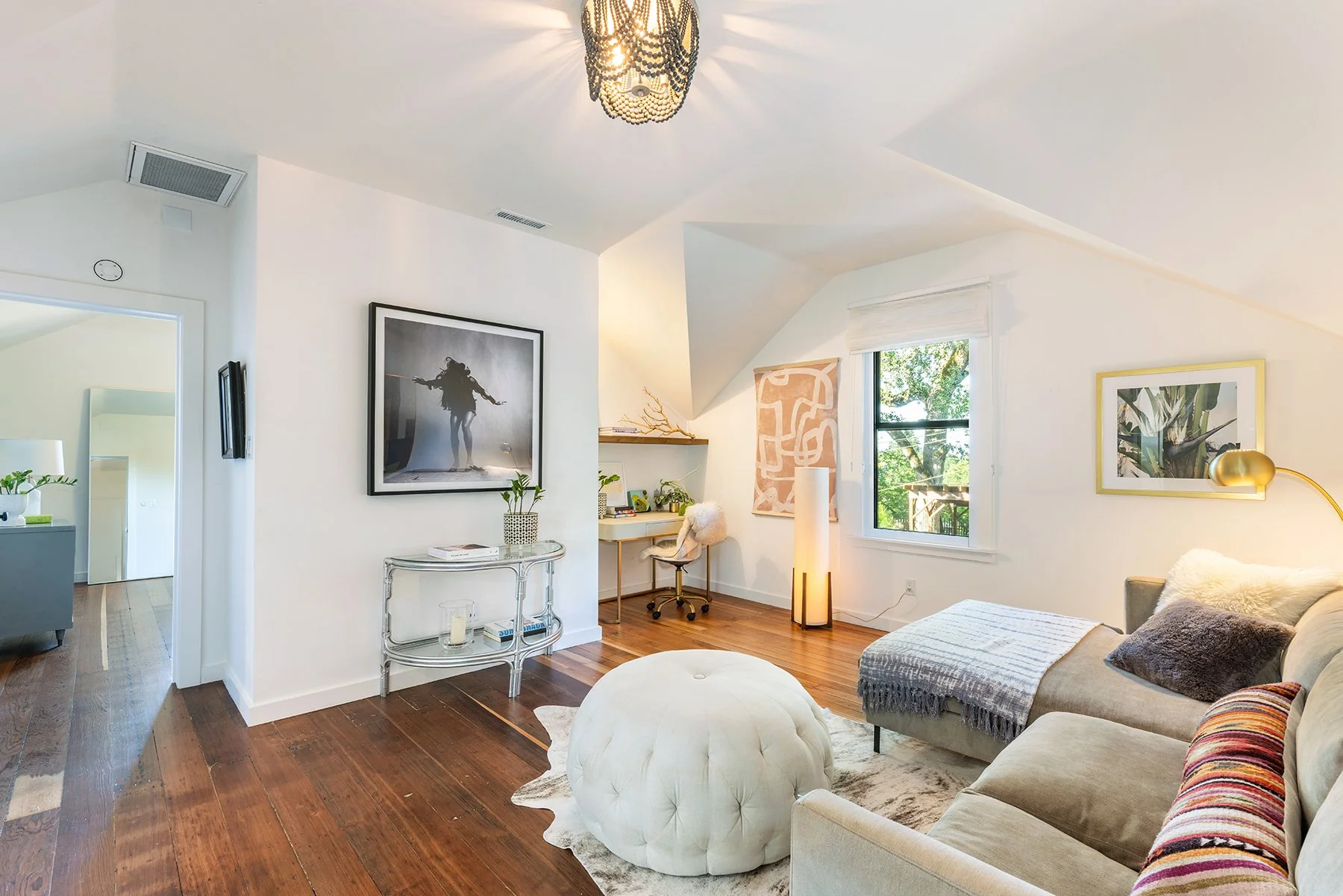 Bright living room with wooden floors, a beige sofa with pillows, a white ottoman, a gold arc floor lamp, wall art, a window with a view of trees, and a small workspace with a desk and chair.